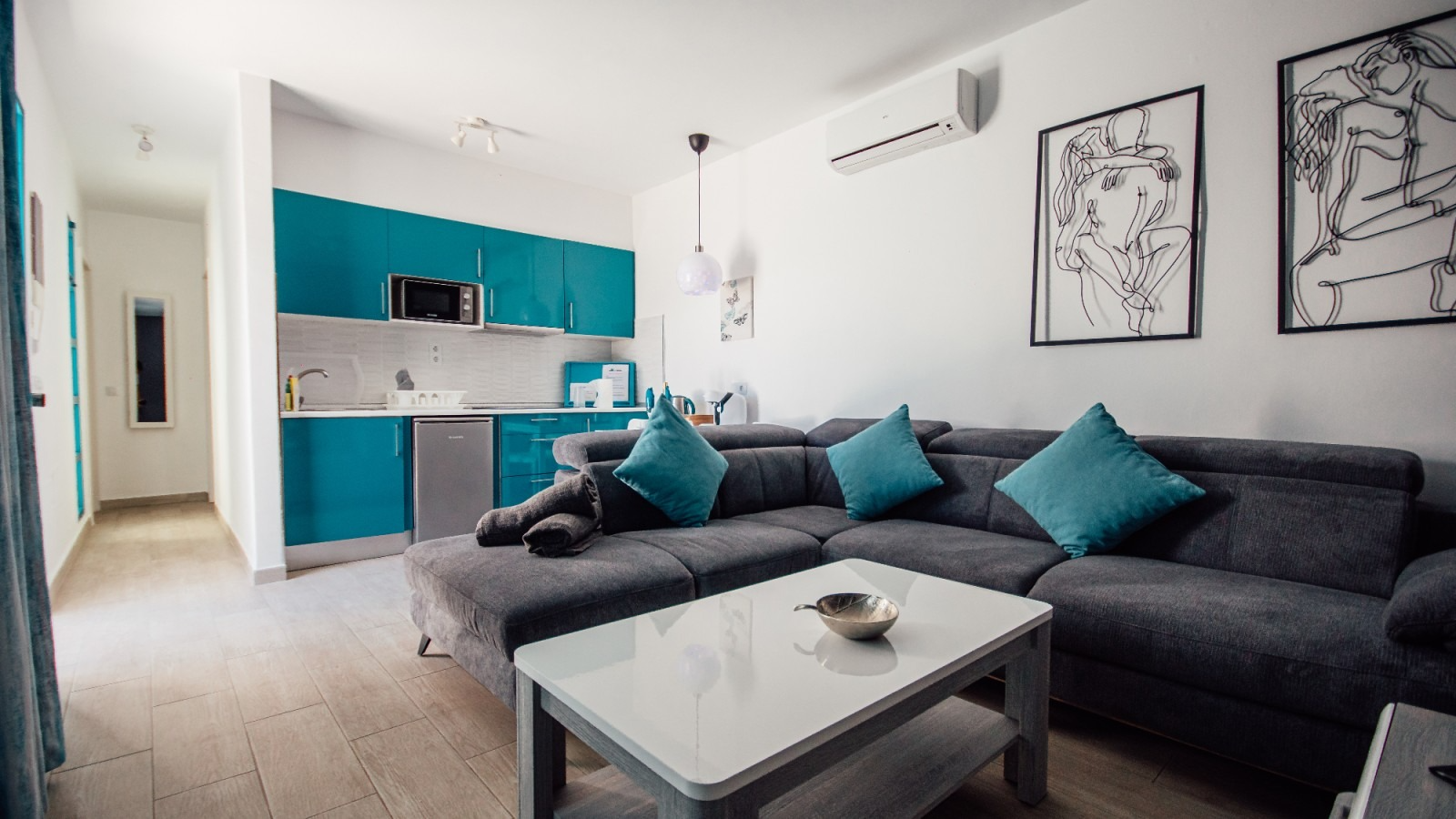 Modern living room with dark gray sectional, teal accents, white coffee table, and open kitchen area