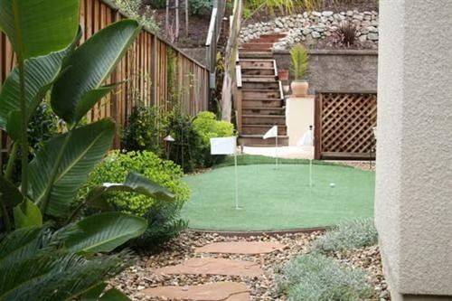 Small backyard mini-golf green with putting hole, surrounded by plants and stone steps.