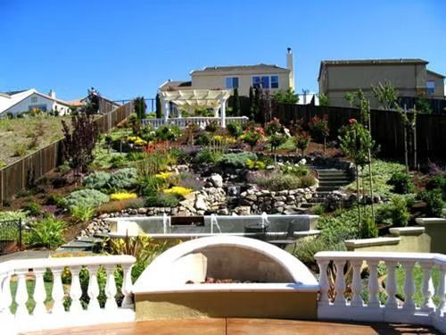 Terraced backyard garden with stone steps, lush plants, and a white balcony railing under a blue sky