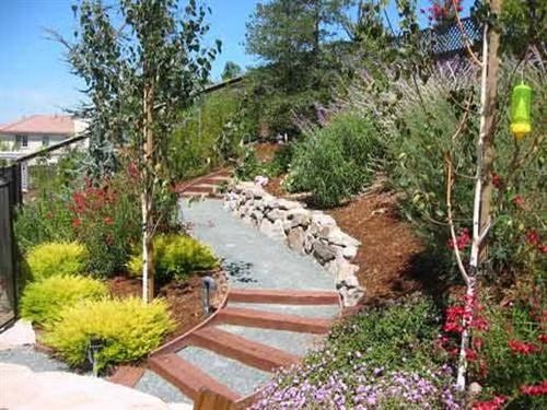 Curved garden path with stone steps, rocks, shrubs, and colorful flowers beside a landscaped hillside.