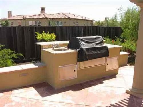 A backyard outdoor kitchen with a stone counter, grill, and pergola, beside a rock garden and a lawn on a sunny day.