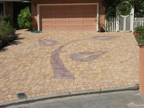 Residential driveway with tan pavers and a garage door, featuring a decorative purple swirl pattern.