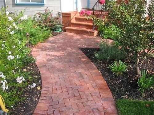 Curved red brick garden path leading to front steps, bordered by green shrubs and flowers.