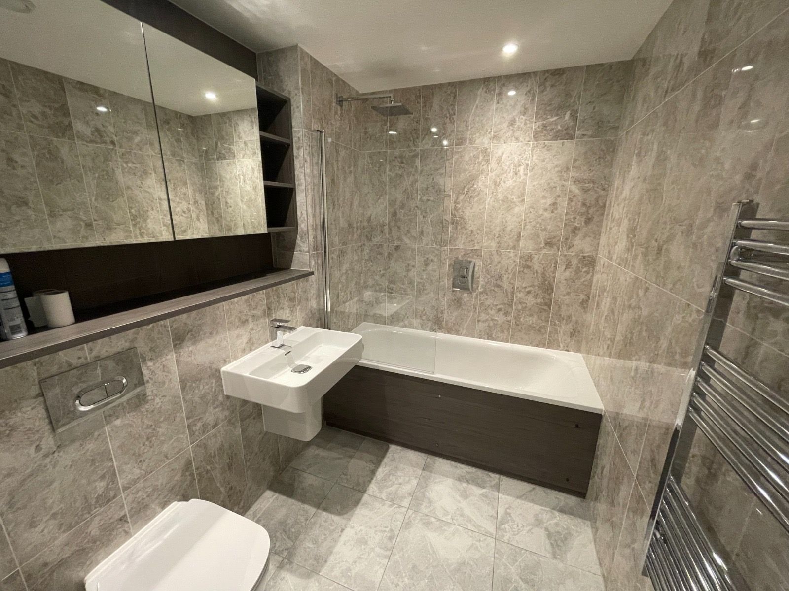 Modern bathroom with gray tile walls, toilet, sink, tub, and towel rack.
