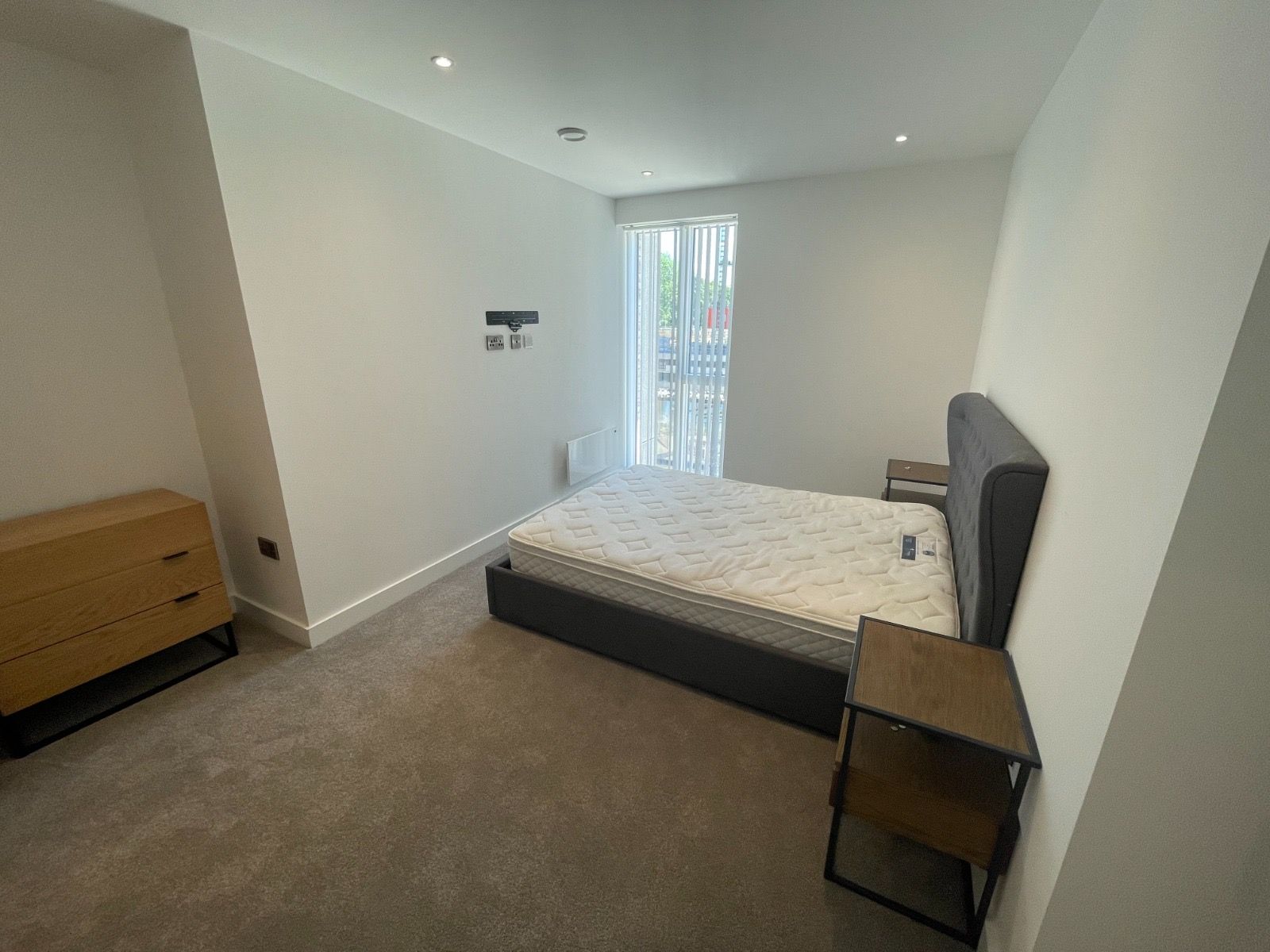 Bedroom with a bed, nightstand, and dresser. White walls, grey carpet. Window with light filtering through.