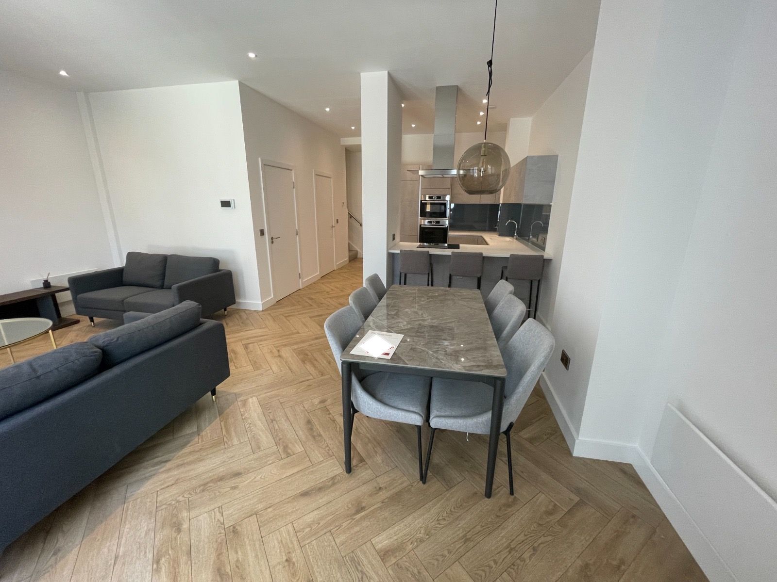 Modern open-plan living space with dining table, kitchen, and two gray sofas on herringbone wood floors.