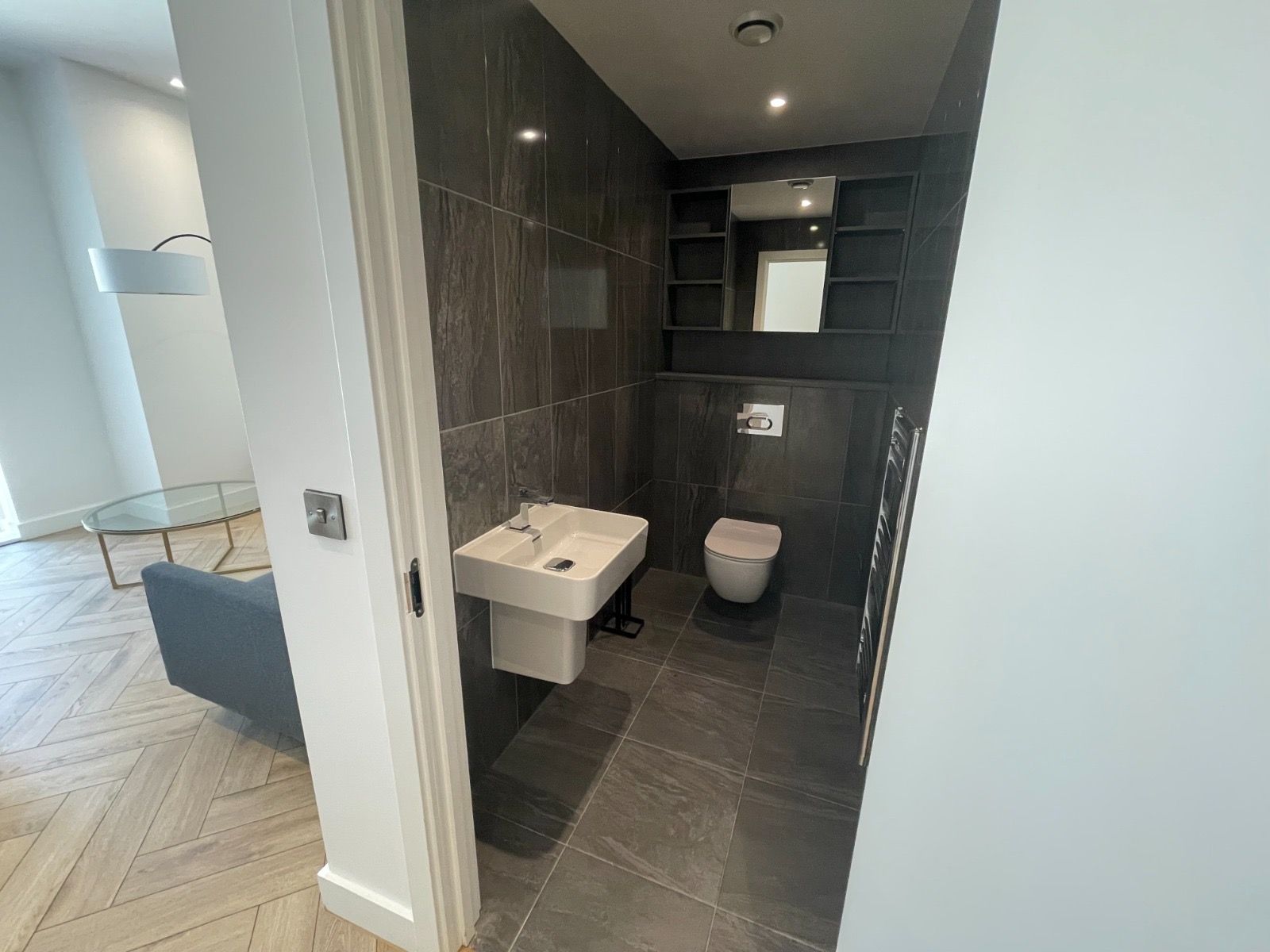 Bathroom with dark tiled walls, toilet, sink, and mirror. View from the doorway into the living space.