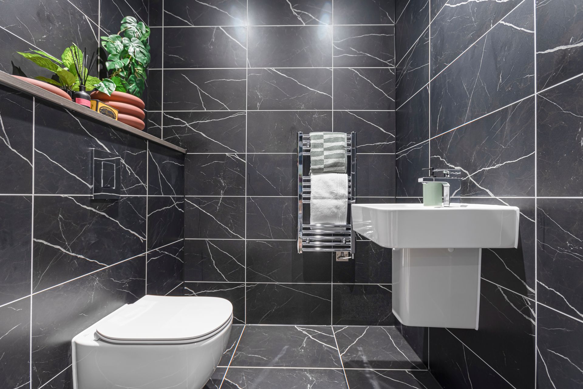 Black tiled bathroom with white pedestal sink, toilet, towel rack, and decorative plants.