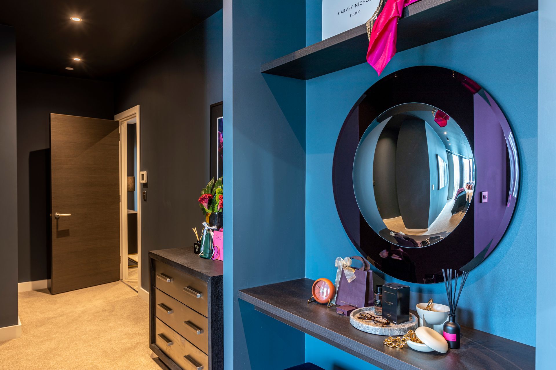 Dressing room with blue walls, round mirror, and a table with accessories. A hallway with a closed door is visible.