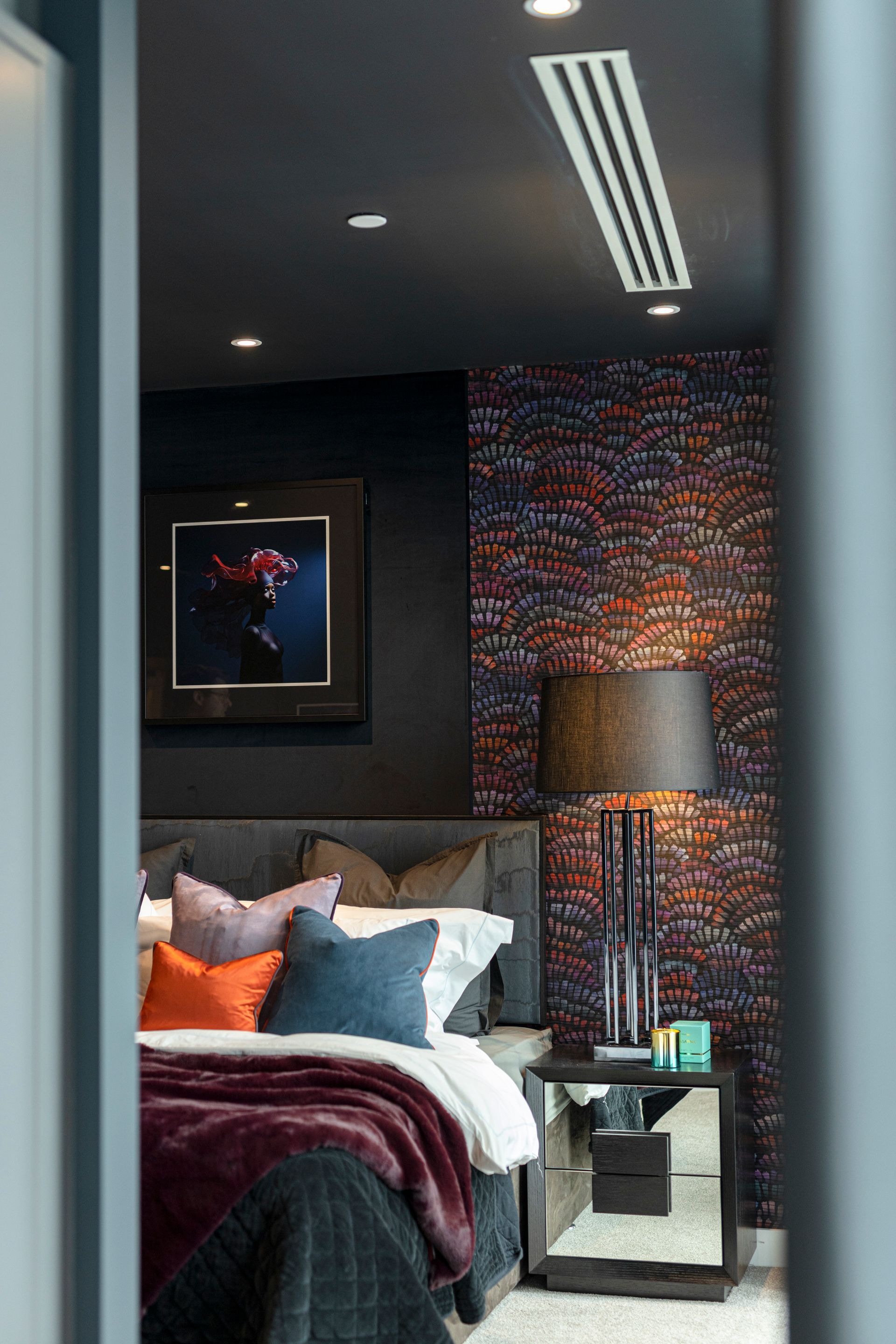 Bedroom with dark walls, patterned wallpaper, a bed with colorful pillows, and a mirrored nightstand.