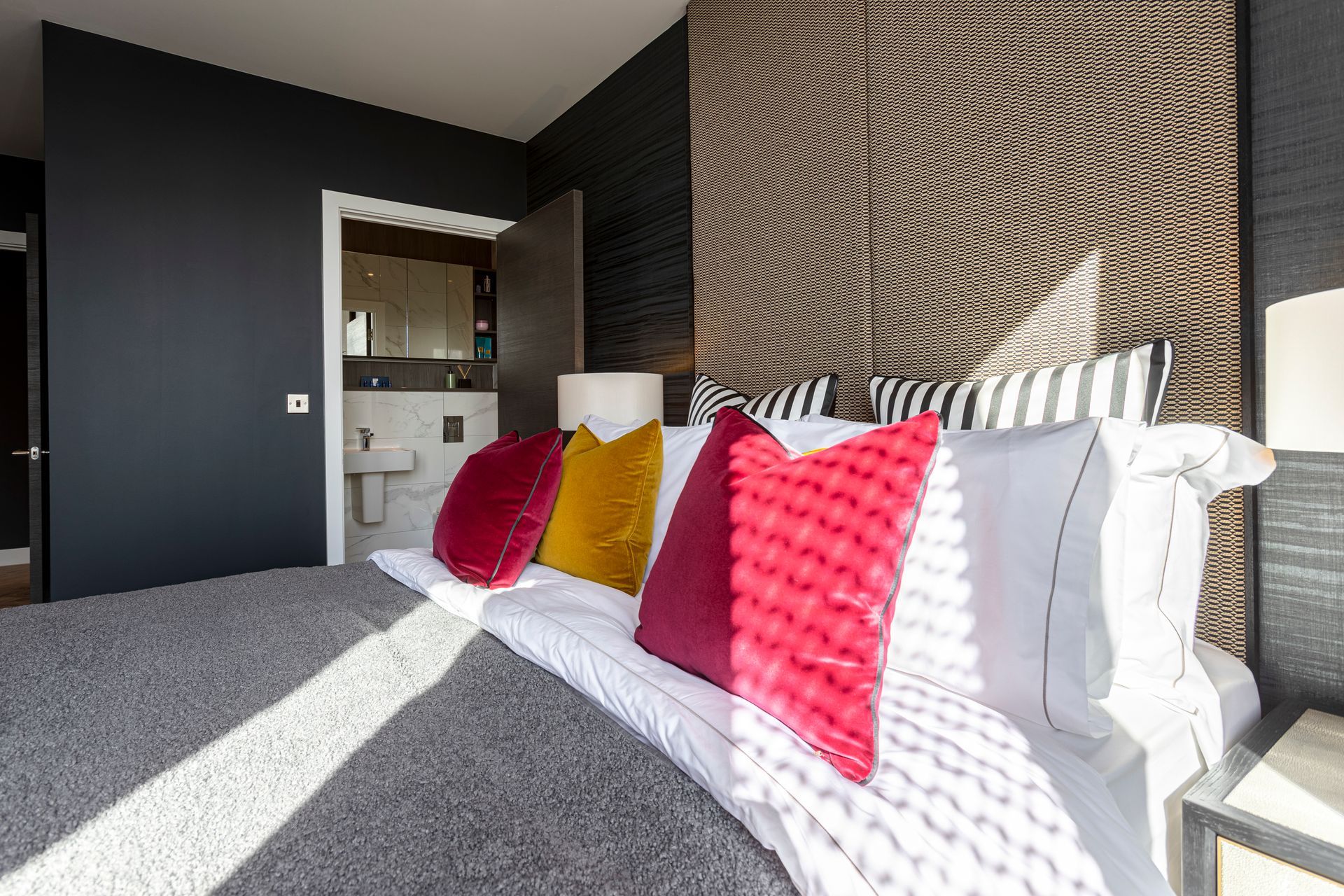 Bedroom with bed, various colored pillows, dark walls, and a view into a bathroom.