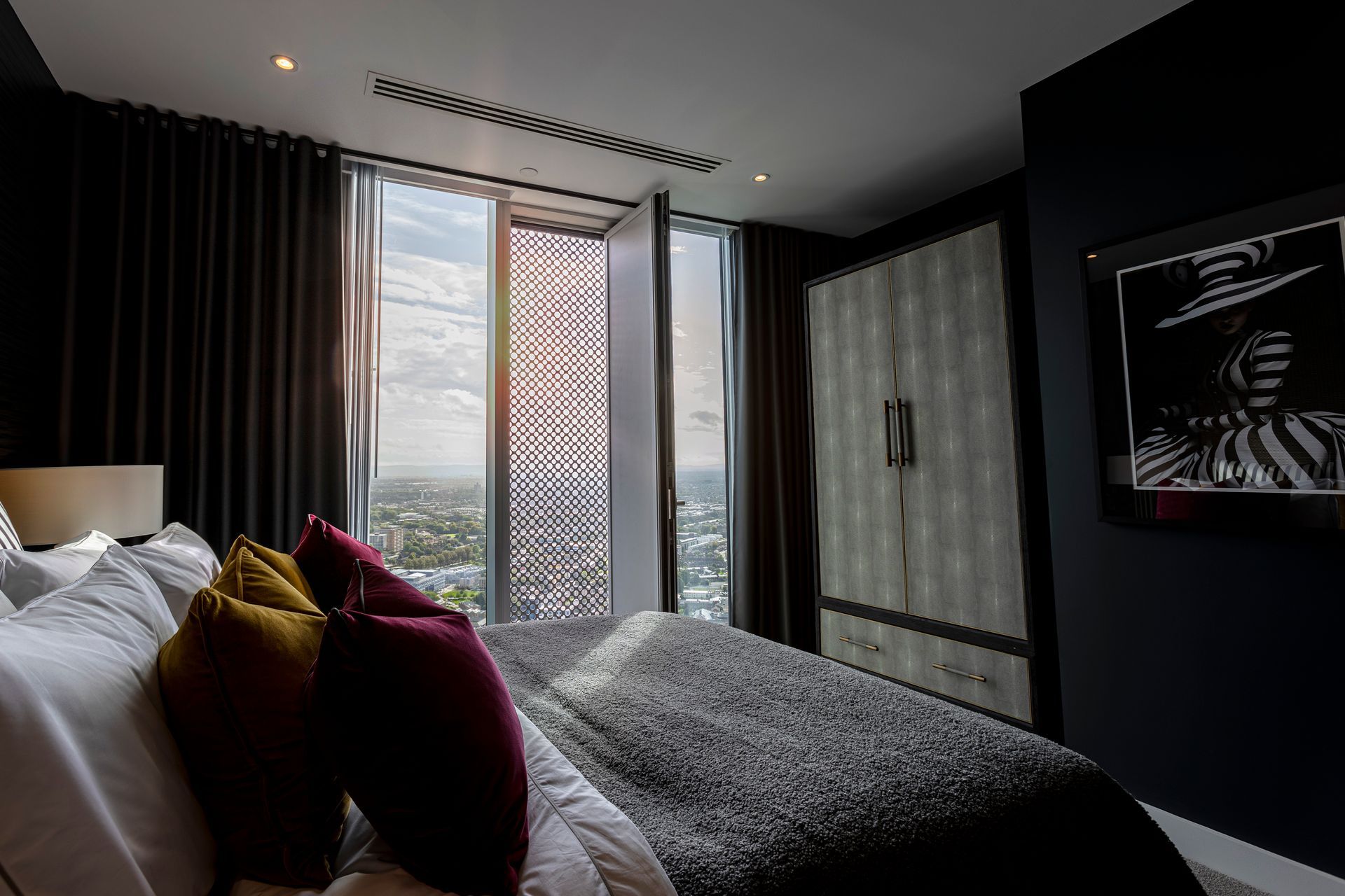 Bedroom with city view, bed with pillows, large windows, patterned decorative panel, dark drapes, wardrobe, artwork.