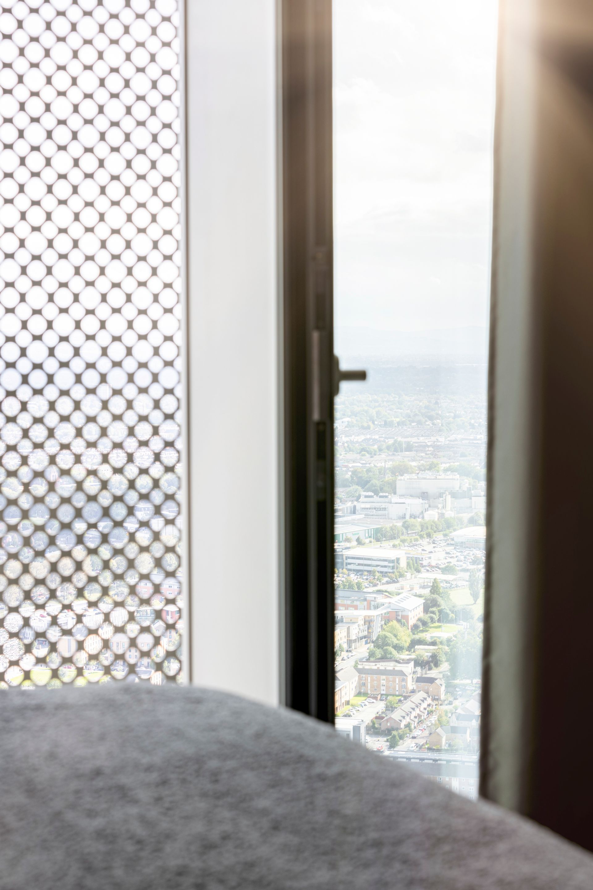Window view overlooking a city. Partially open window reveals grid pattern screen, city buildings, and hazy sky.