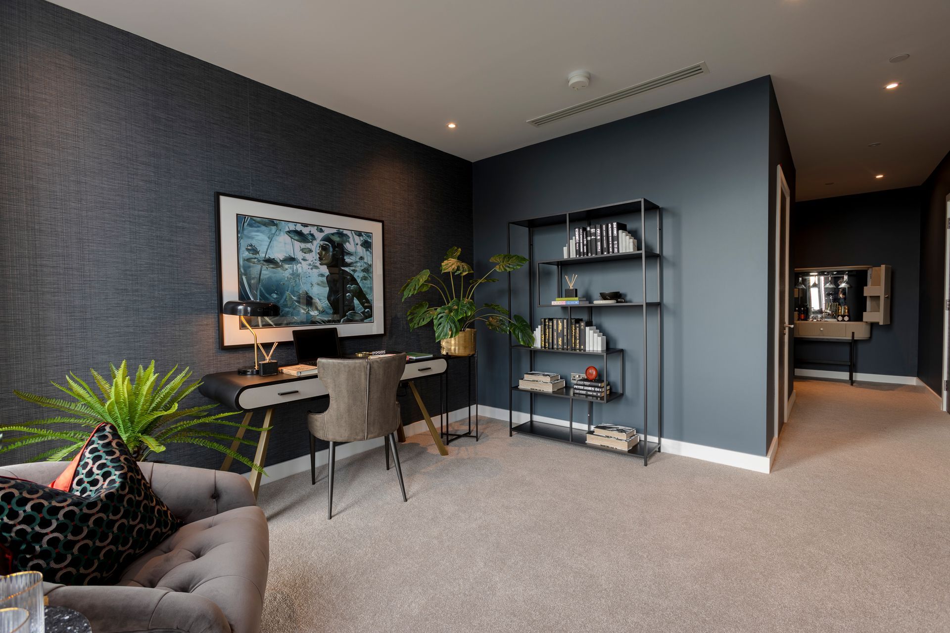 Home office with desk, bookshelf, and a gray sofa. Dark blue walls, carpet floor.