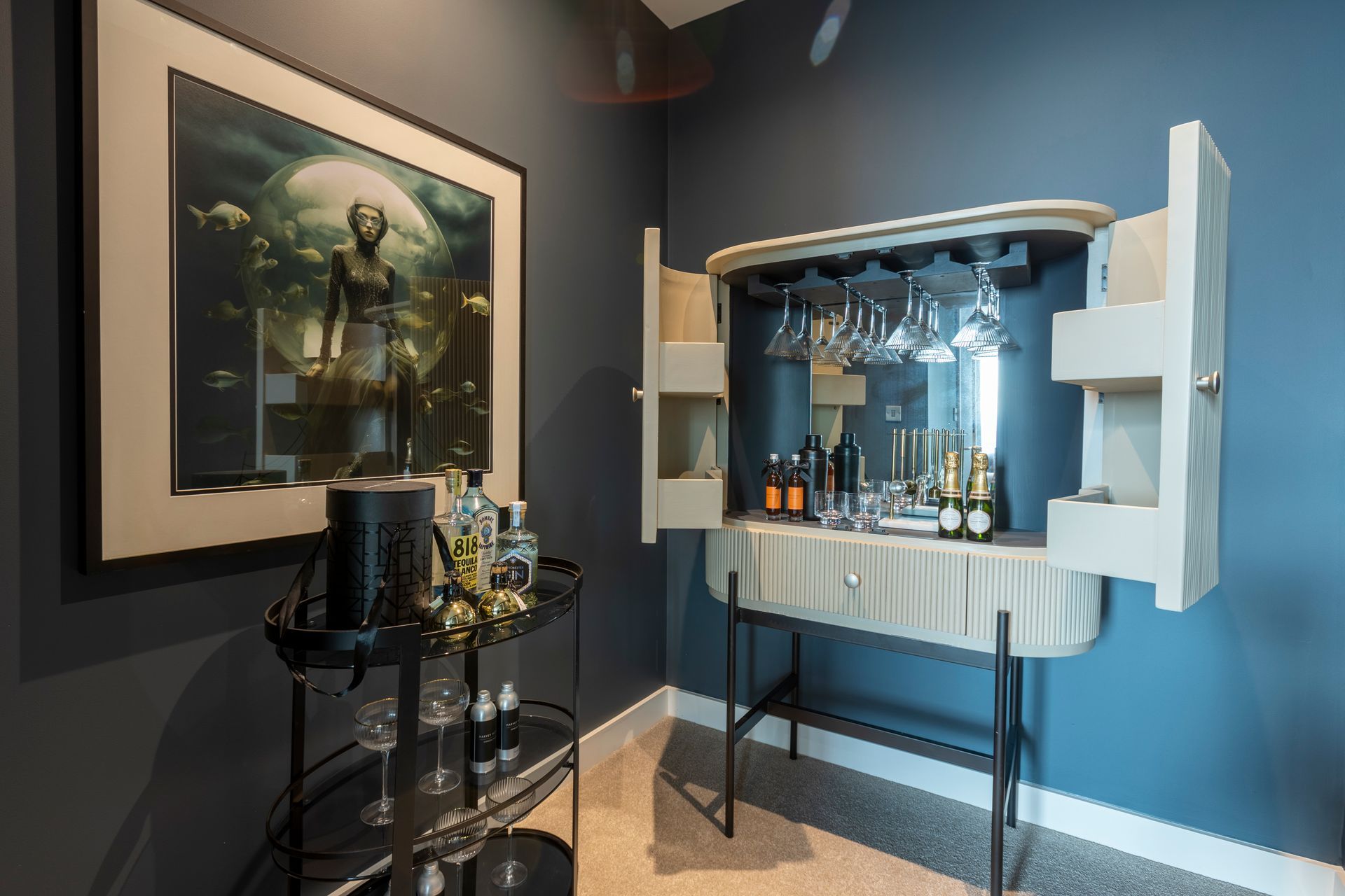 A bar cabinet open against a blue wall next to artwork and a black bar cart.