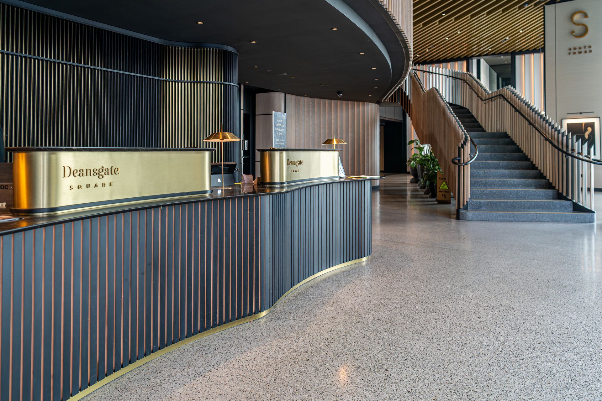 Hotel reception area with curved front desk and staircase. Gold accents, dark gray and wood tones.