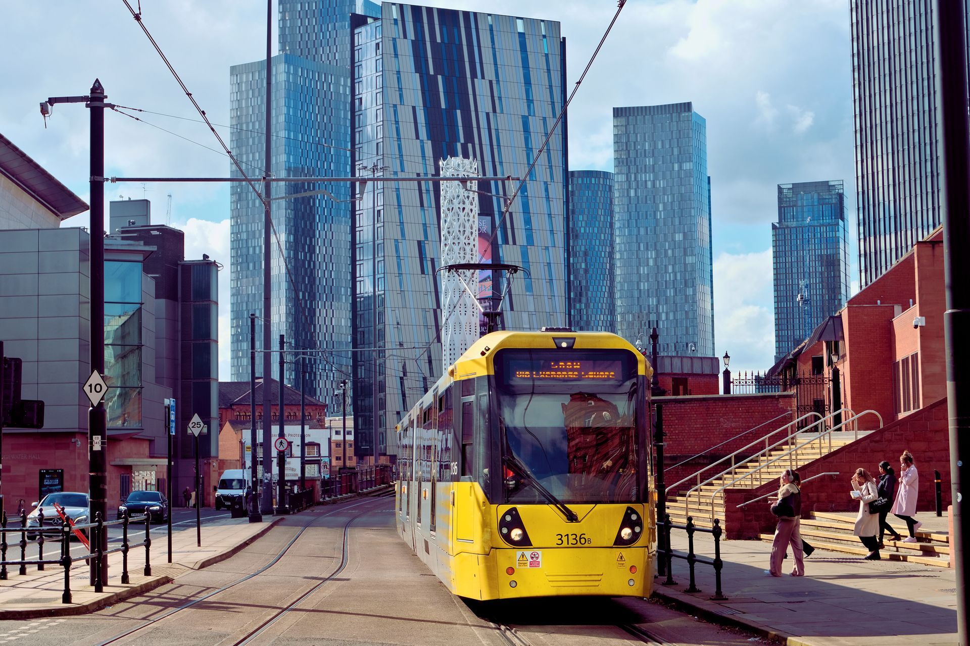Yellow tram on tracks, tall modern buildings in background, pedestrians on stairs.