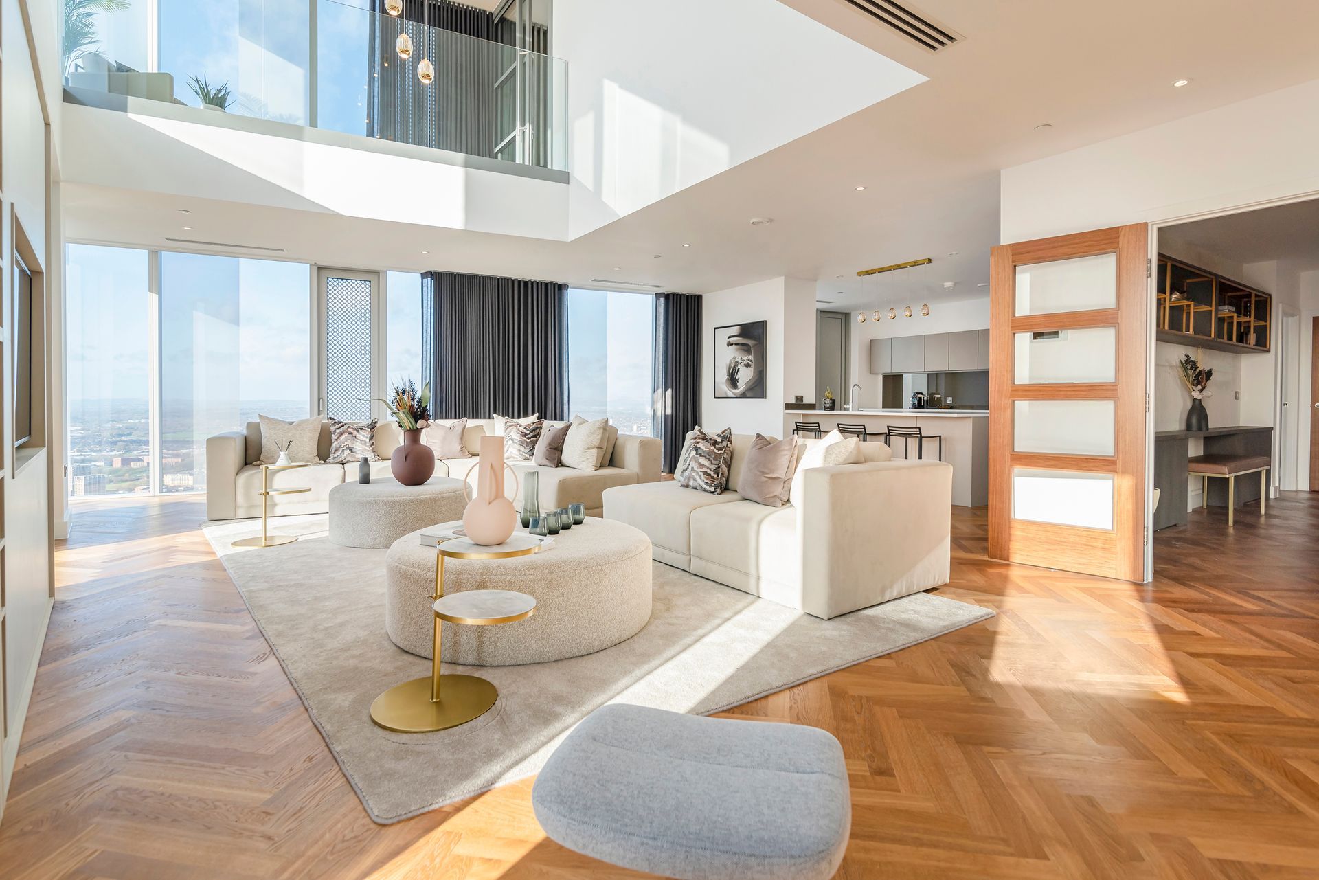 Modern living room with light-colored sofas, circular ottoman, and city views. Wooden floors and open kitchen area.
