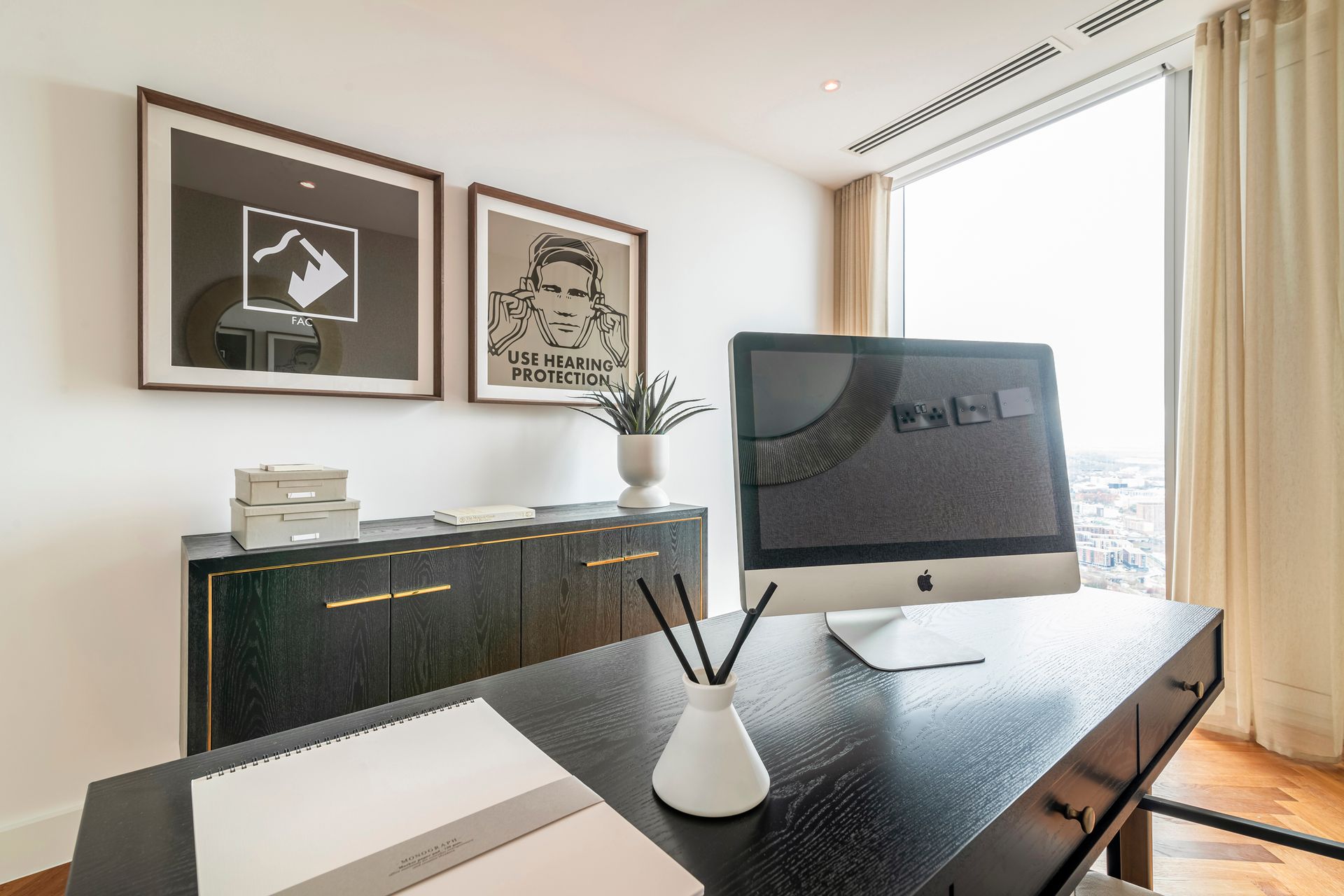 Office with black desk, large computer, art, and sideboard near a window with curtains.