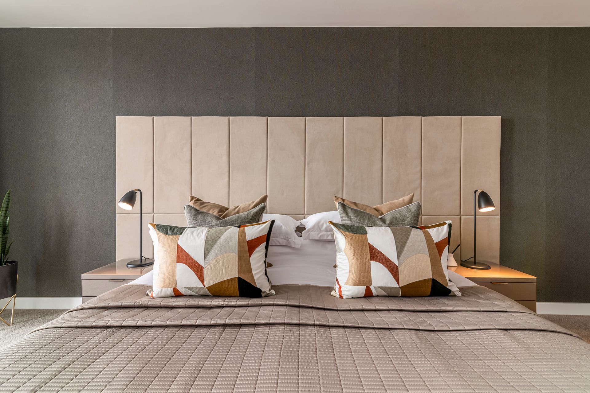 Bedroom with beige upholstered headboard, lamps, patterned pillows, bedspread, and dark gray textured wall.