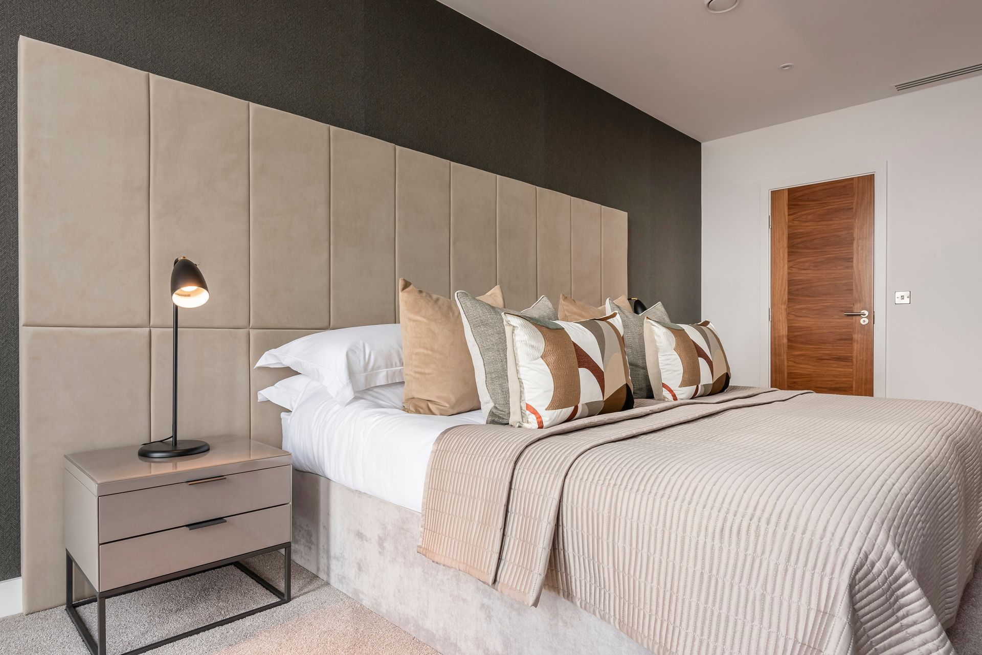 Bedroom with large padded headboard, bed with decorative pillows, nightstand with lamp, and door.