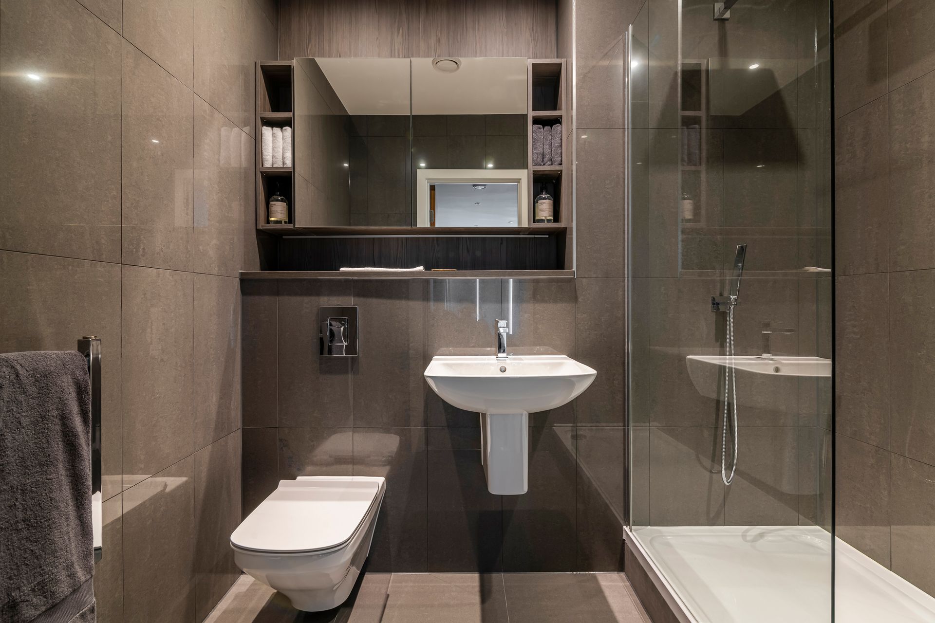Modern bathroom with gray tiles, toilet, sink, mirror, and glass shower.