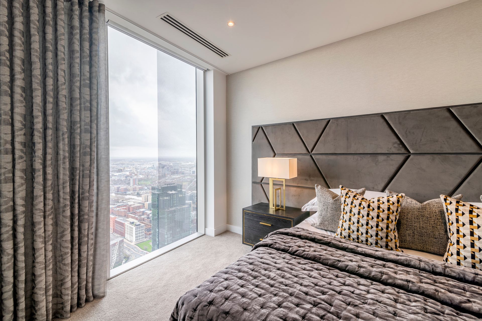 Bedroom with large window overlooking city. Gray and gold accents, plush bed, and decorative pillows.