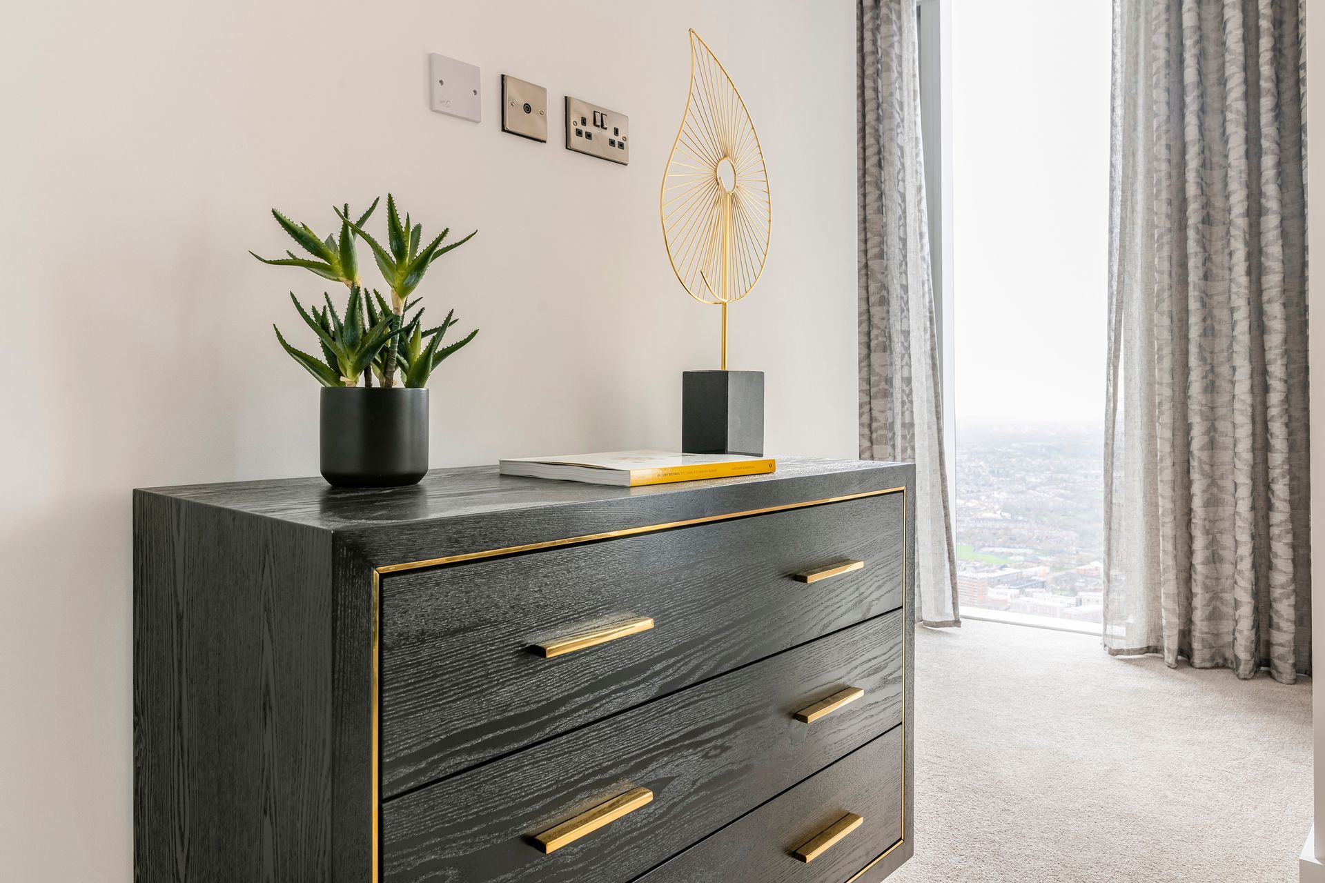 Black dresser with gold accents, plants, and art in a modern room with a window and sheer curtains.