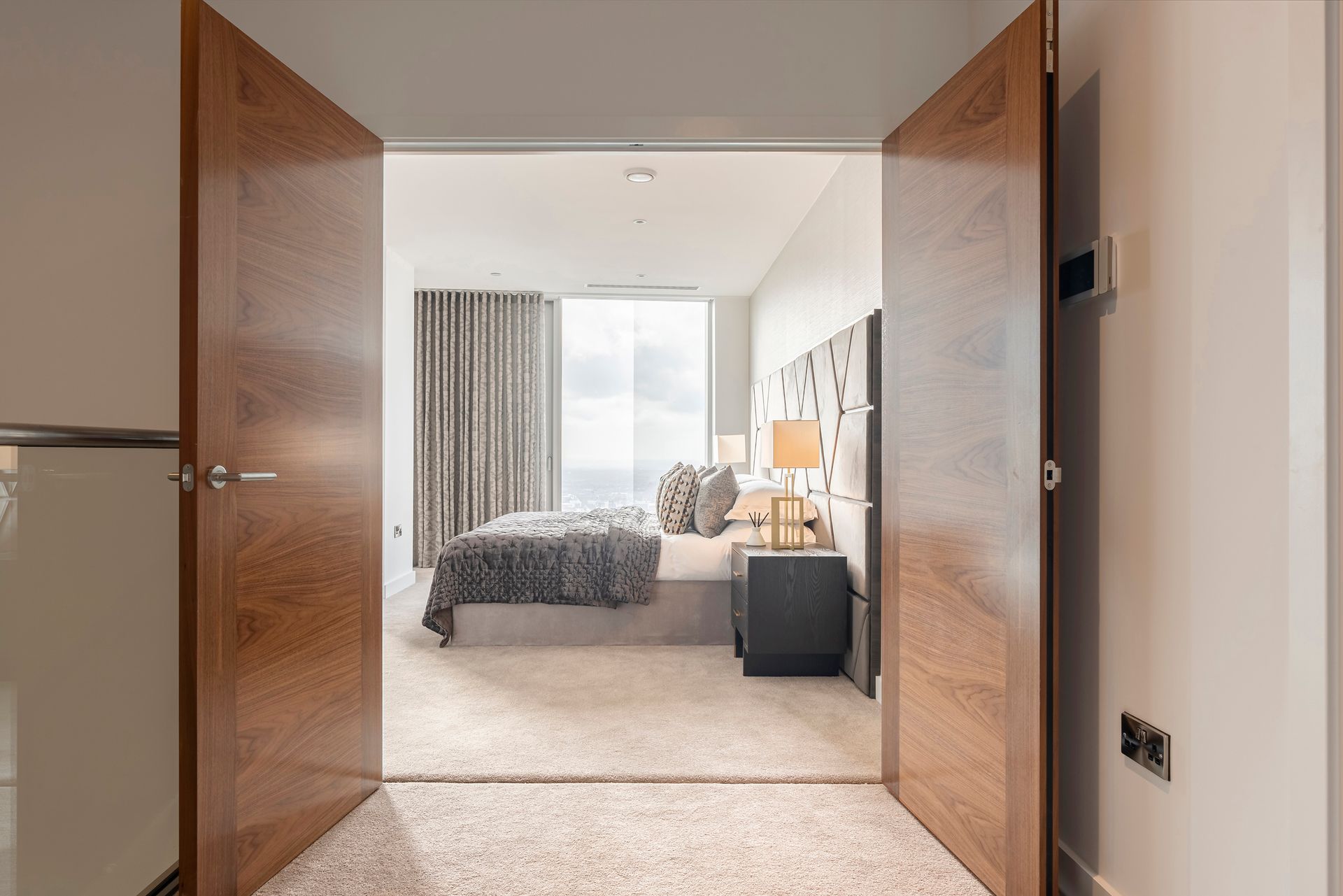 Bedroom interior seen through open wooden doors. Neutral colors, bed, nightstand, and window with curtains.