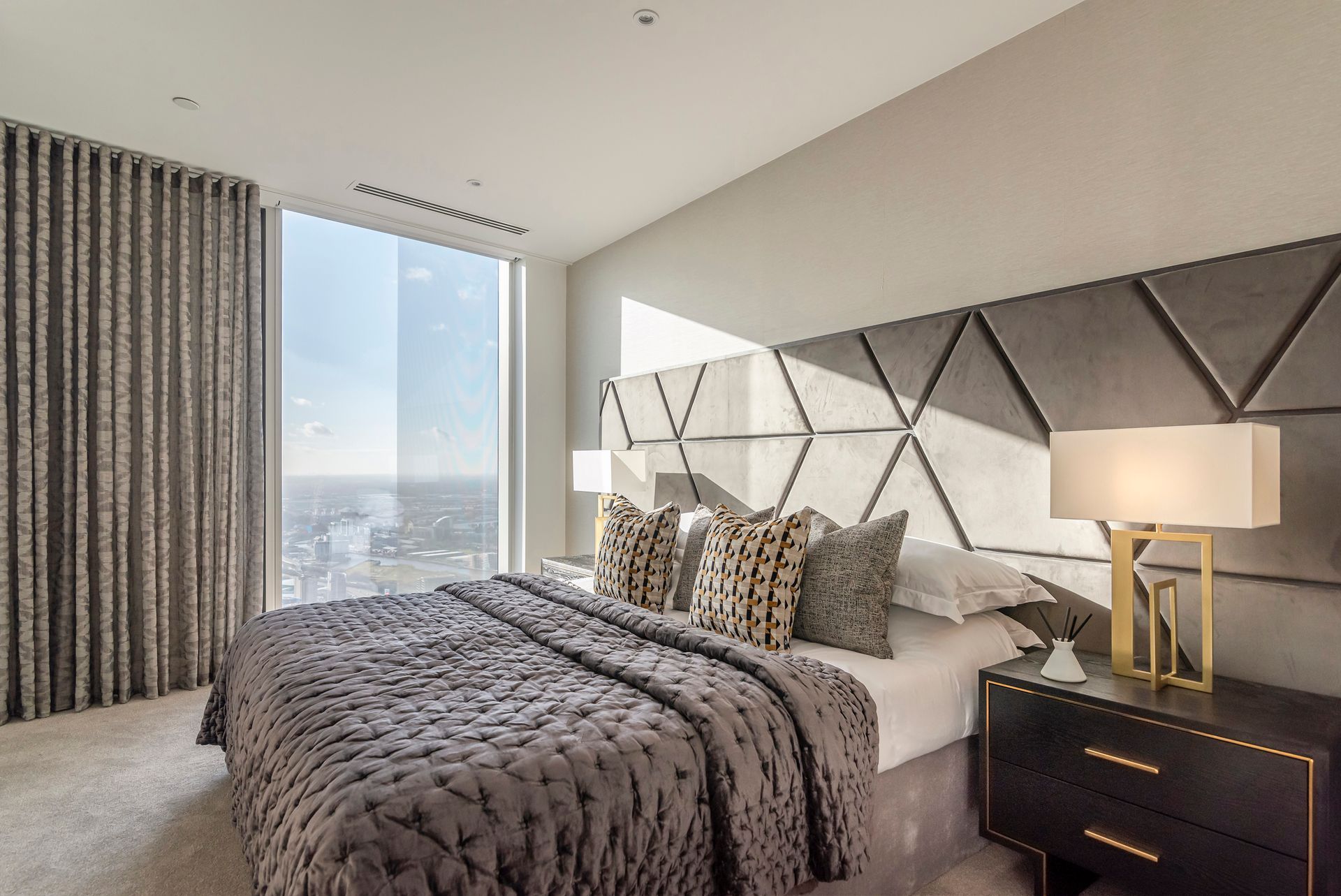 Bedroom with large window, gray bedspread, geometric headboard, and gold bedside lamps.