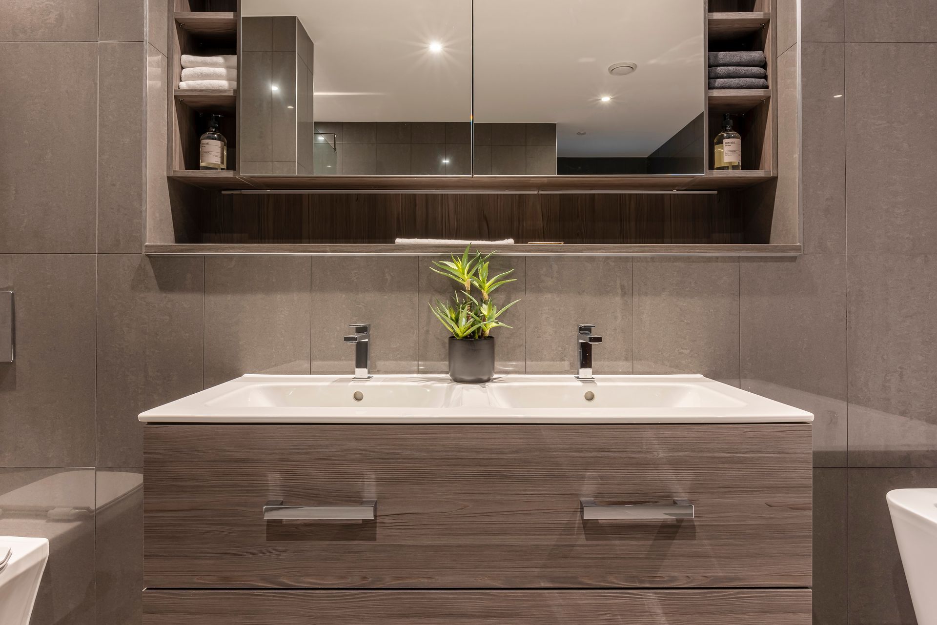 Bathroom vanity with double sinks, gray tiled wall, dark wood cabinet, mirror and shelving.