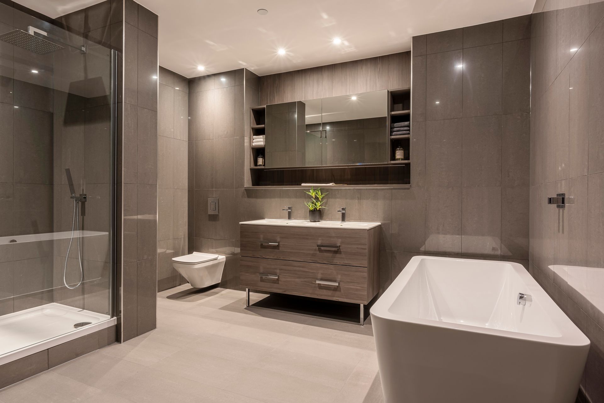 Modern bathroom with gray tiled walls and flooring, a glass shower, toilet, wooden vanity, and a white soaking tub.