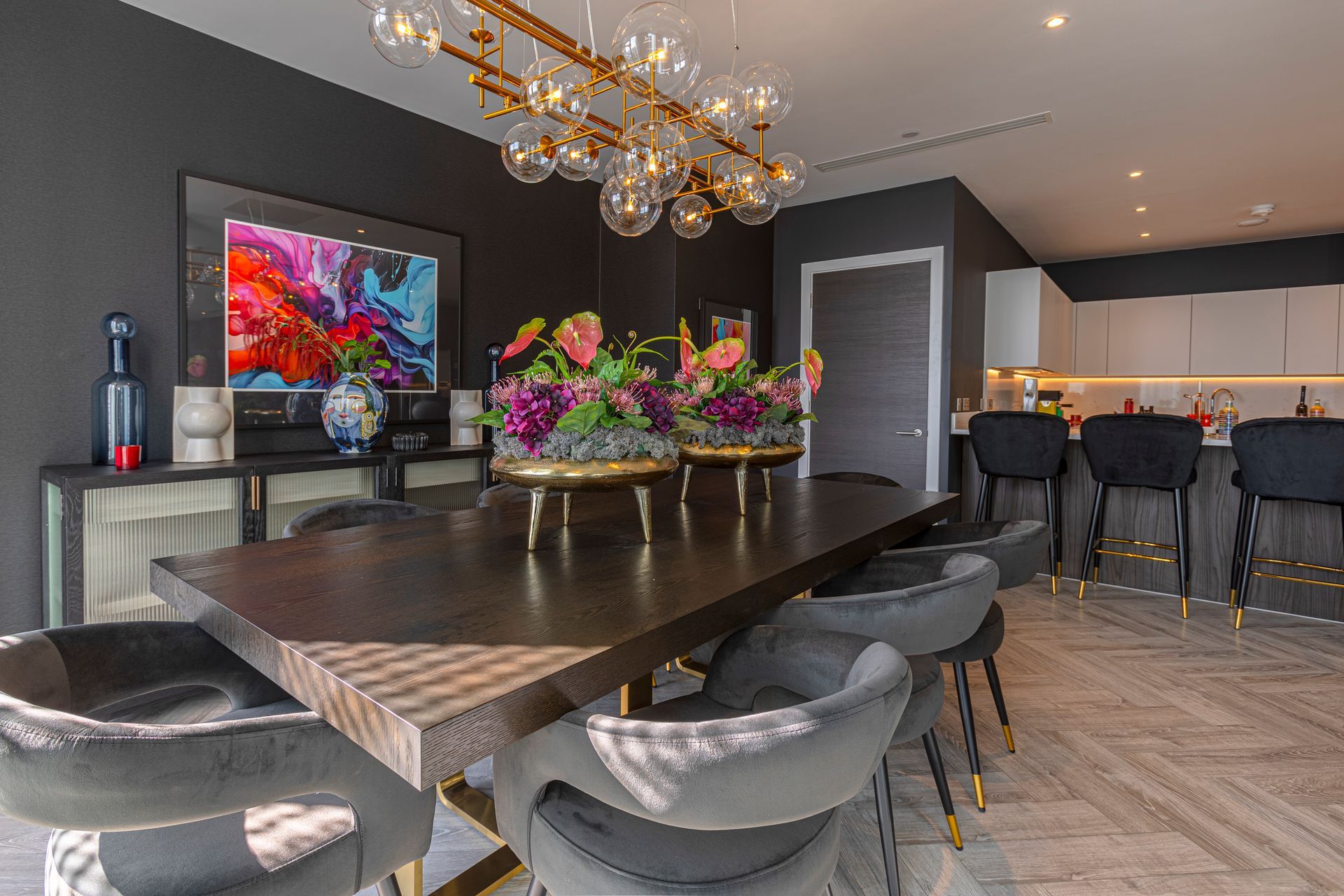 Dark dining room with large wooden table, gray chairs, gold chandelier, and colorful artwork.