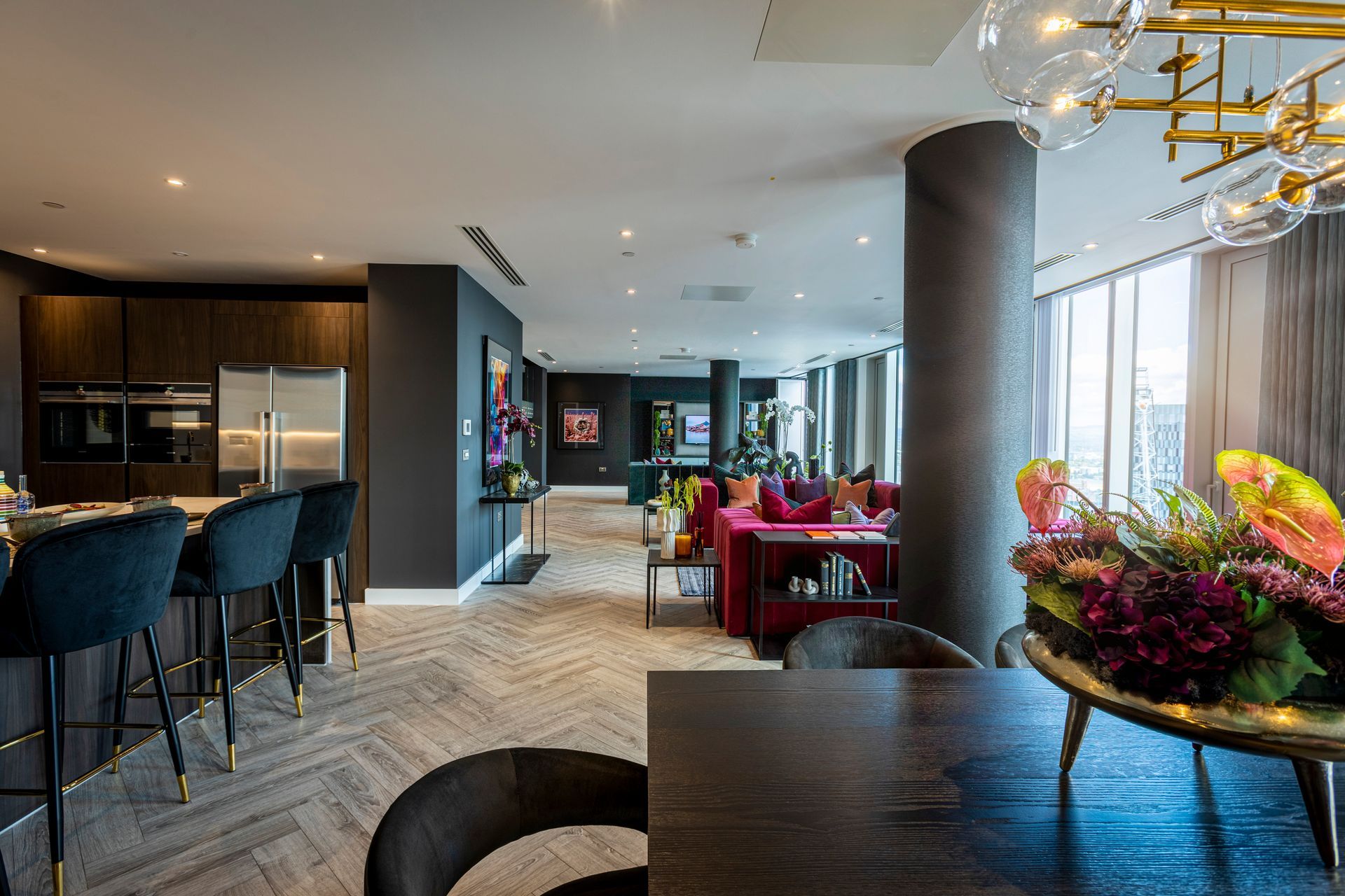 Modern open-plan living space with kitchen, dining, and seating areas. Dark tones, city view, floral centerpiece.
