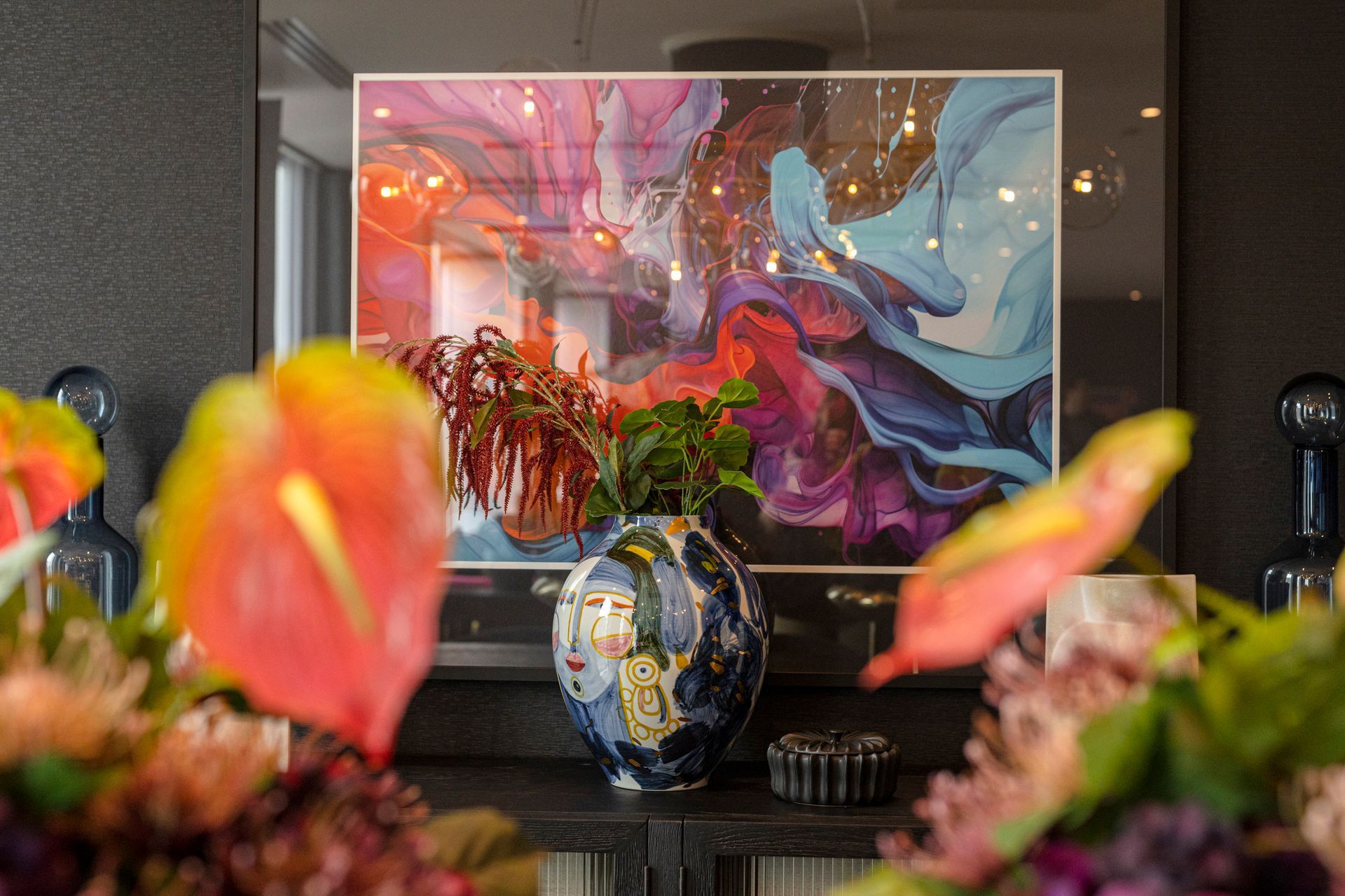 Abstract art framed above a vase of flowers, with colorful blooms and dark background.