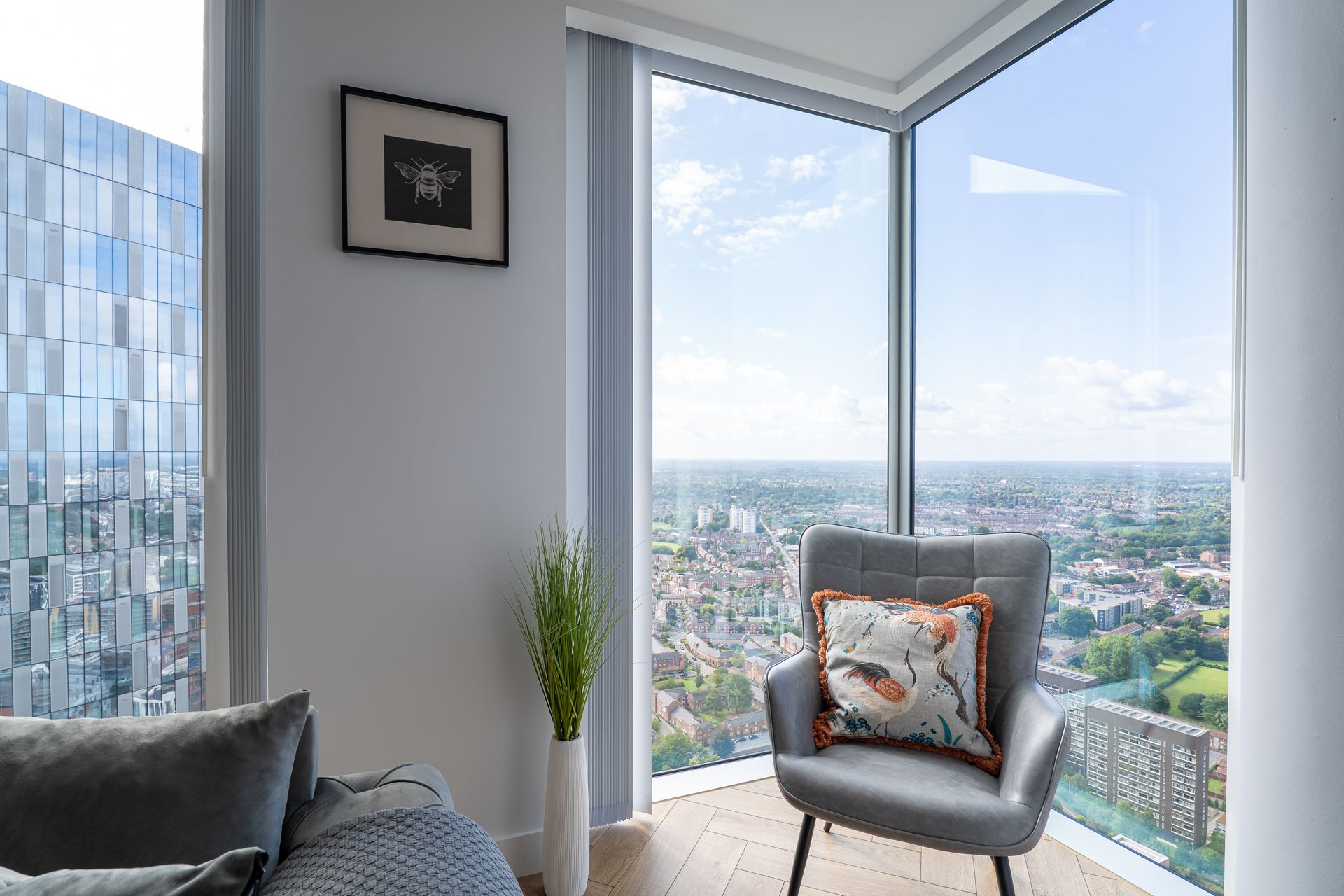Interior with city view, grey armchair, art, large windows, and vase with greenery.