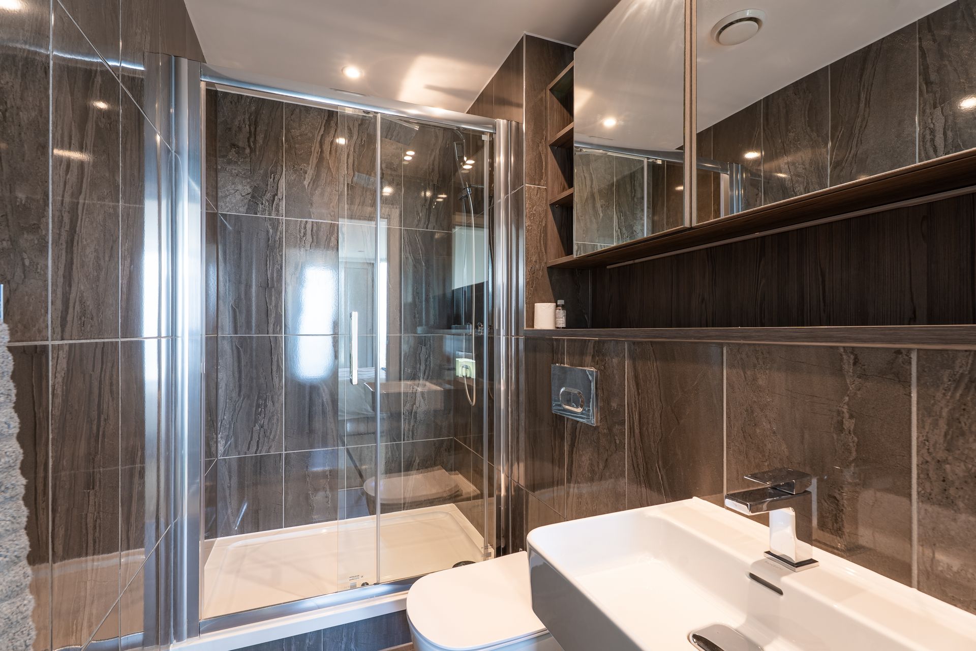 Modern bathroom with a glass shower, white sink, and dark gray tiled walls.