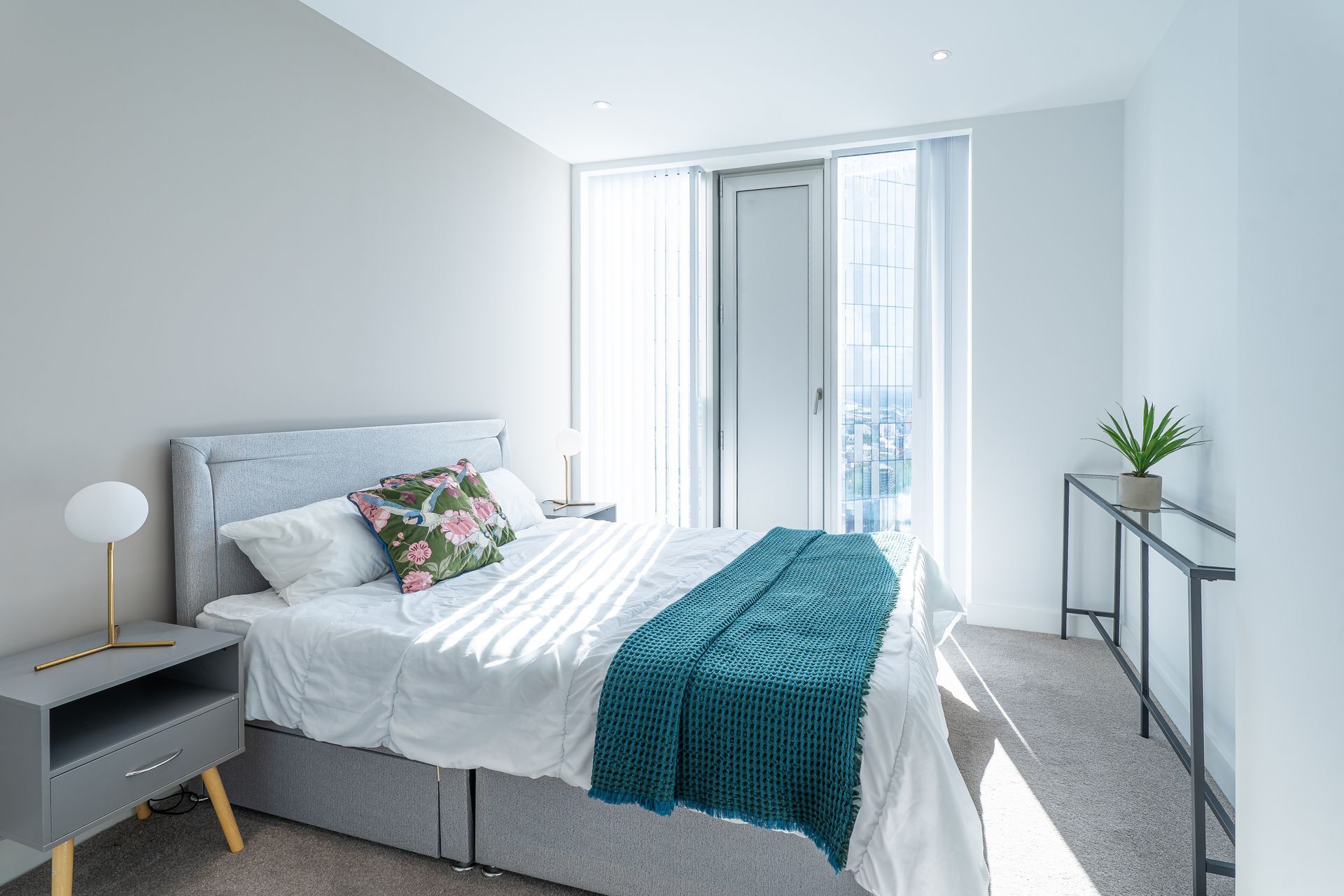 Bedroom with a gray bed, teal blanket, and a sliding door. A small table with a plant is on the right.