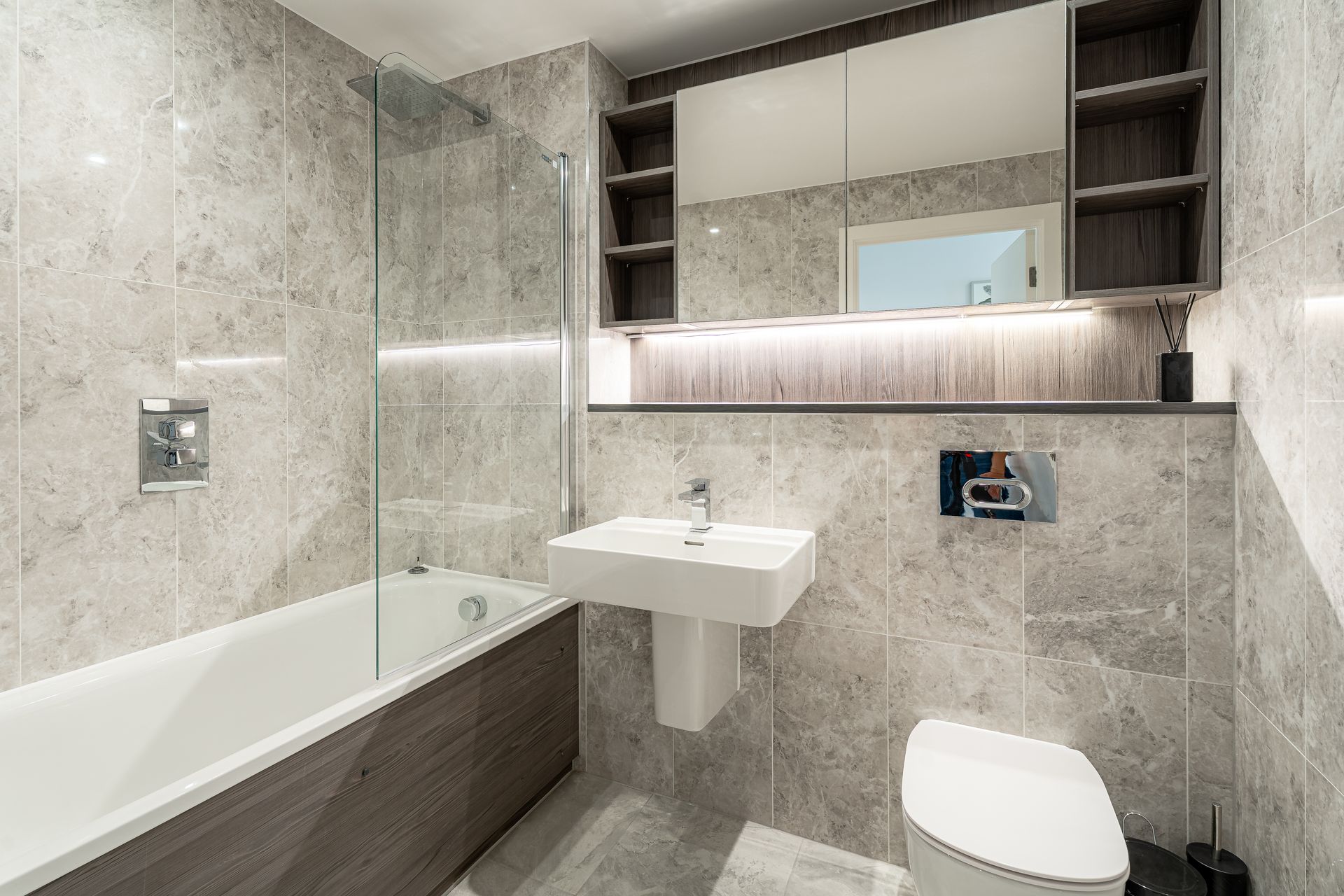 Modern bathroom with gray tile, bathtub, sink, toilet, and mirror cabinet with shelves.