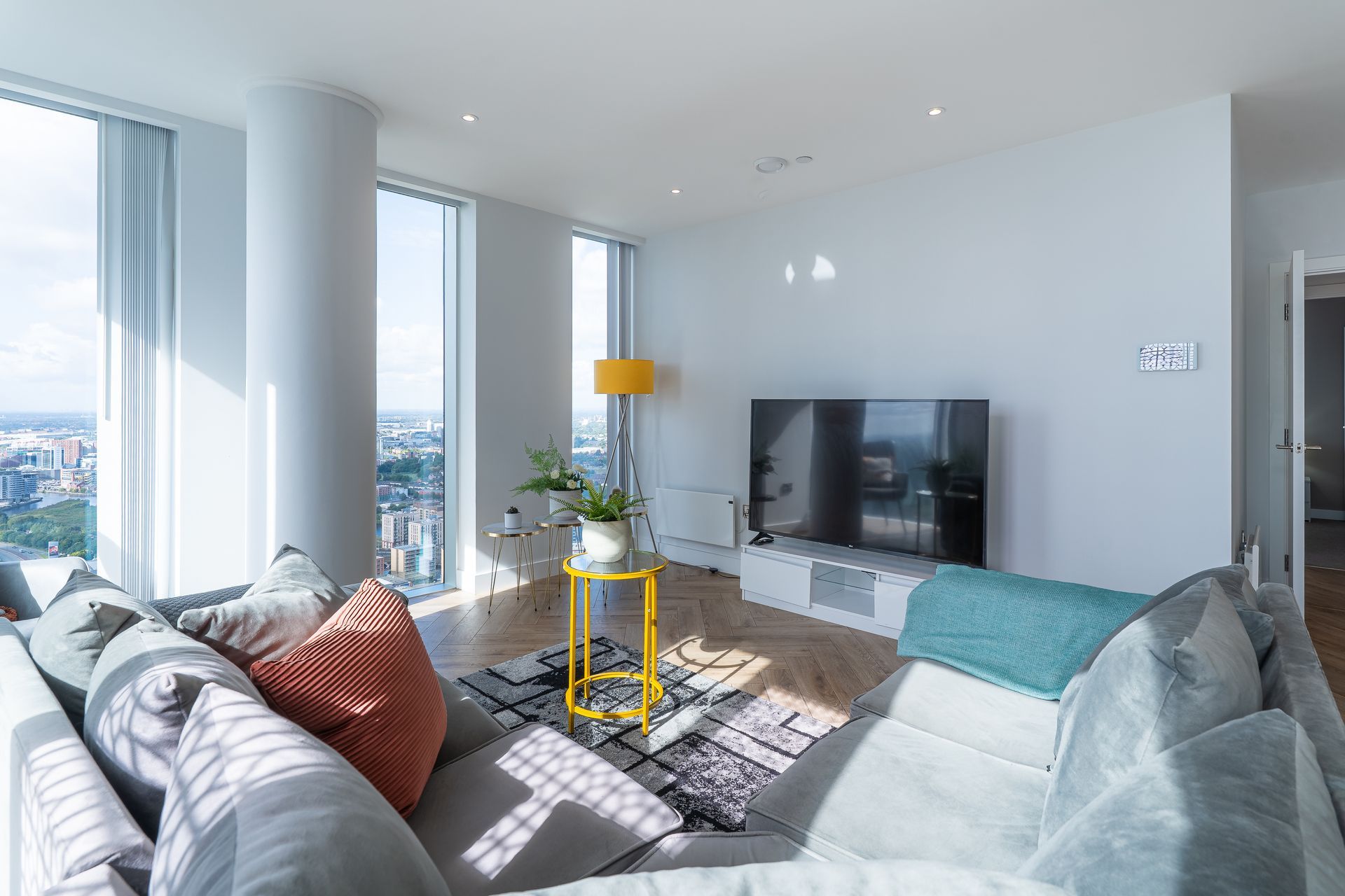 Living room with large windows, gray sectional sofa, TV, yellow accents, and city view.
