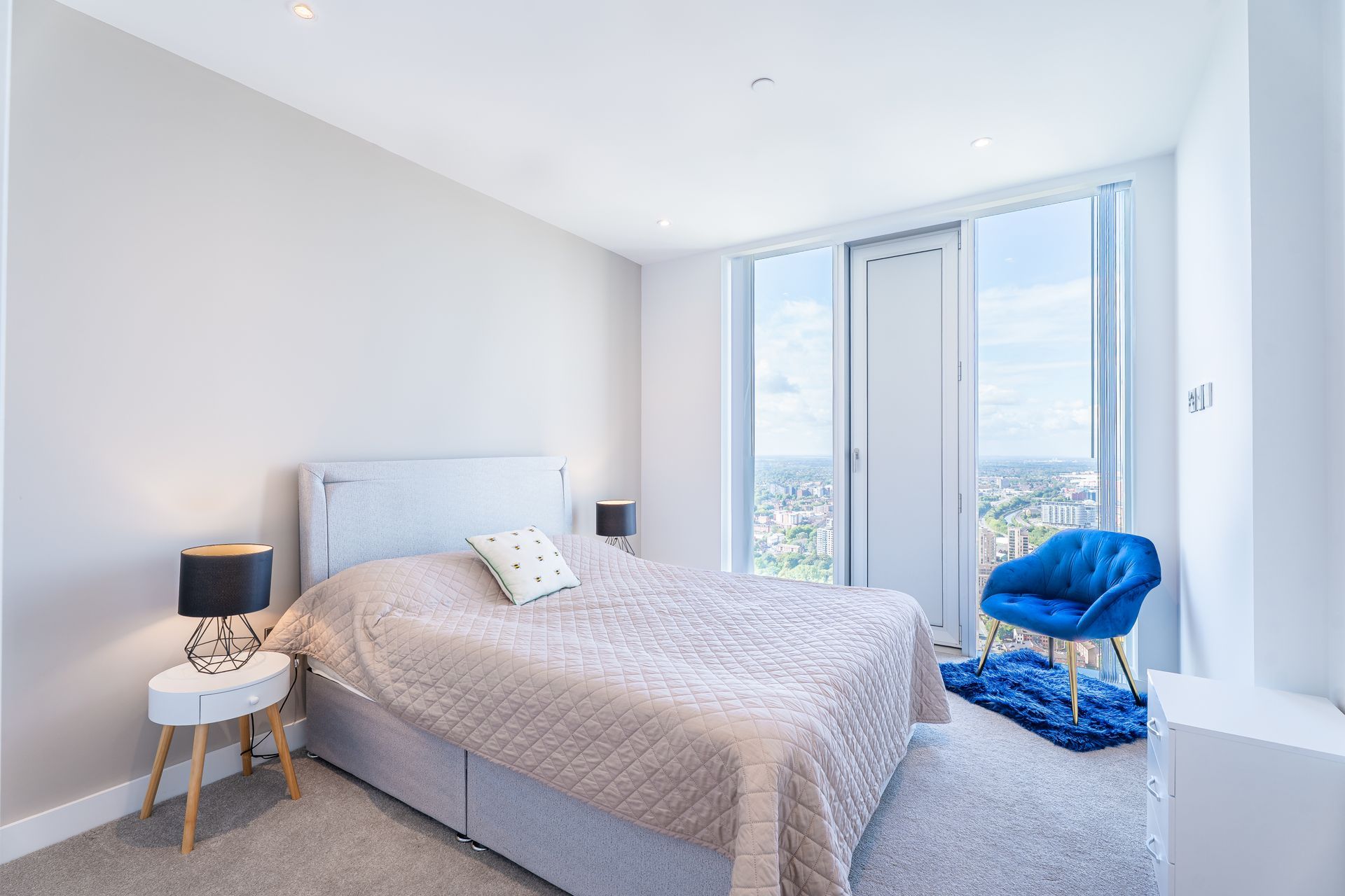 Bedroom with a large bed, nightstands, and a blue chair. Large windows offer a city view.
