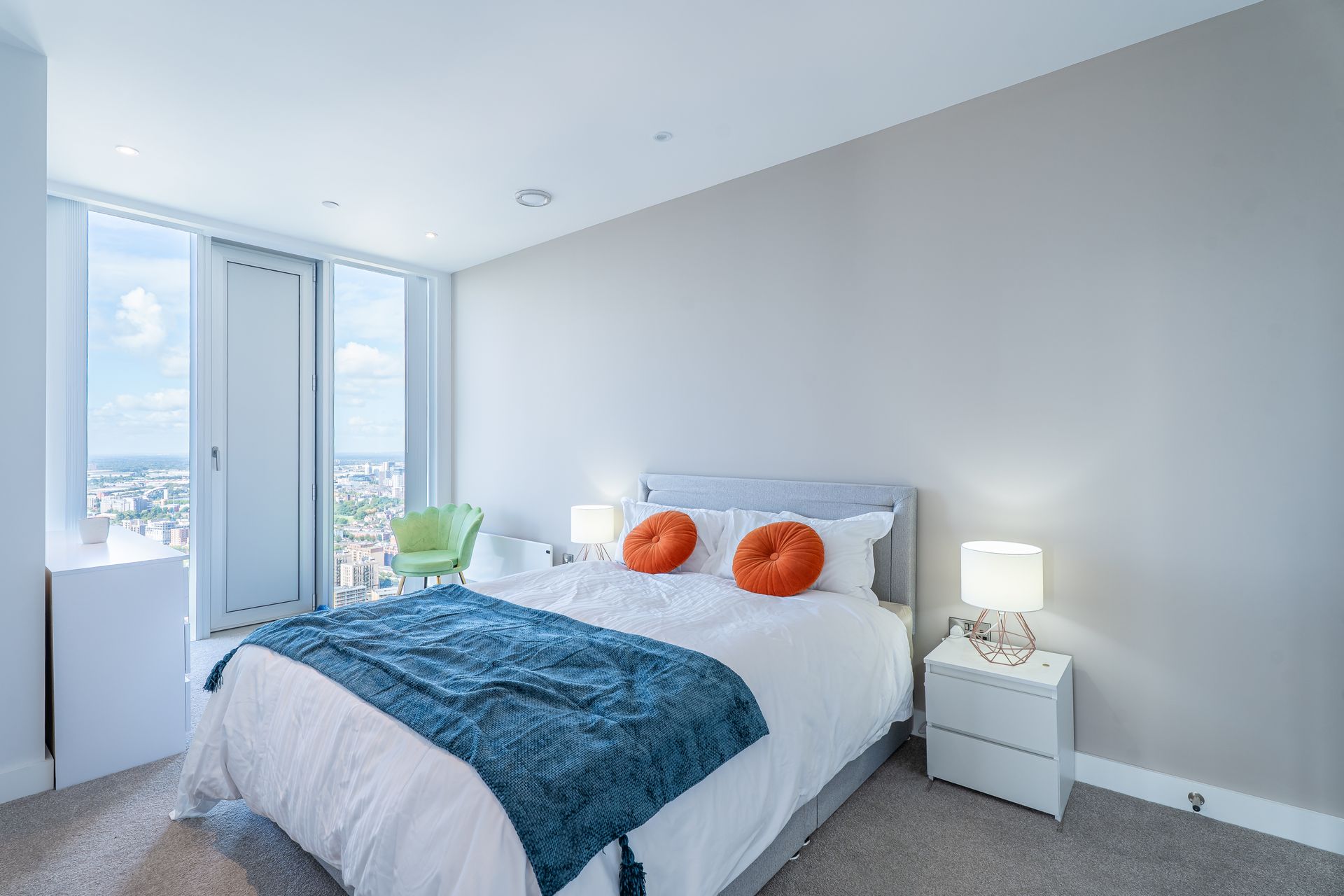 Bedroom with bed, orange pillows, blue throw, bedside tables, and a city view.