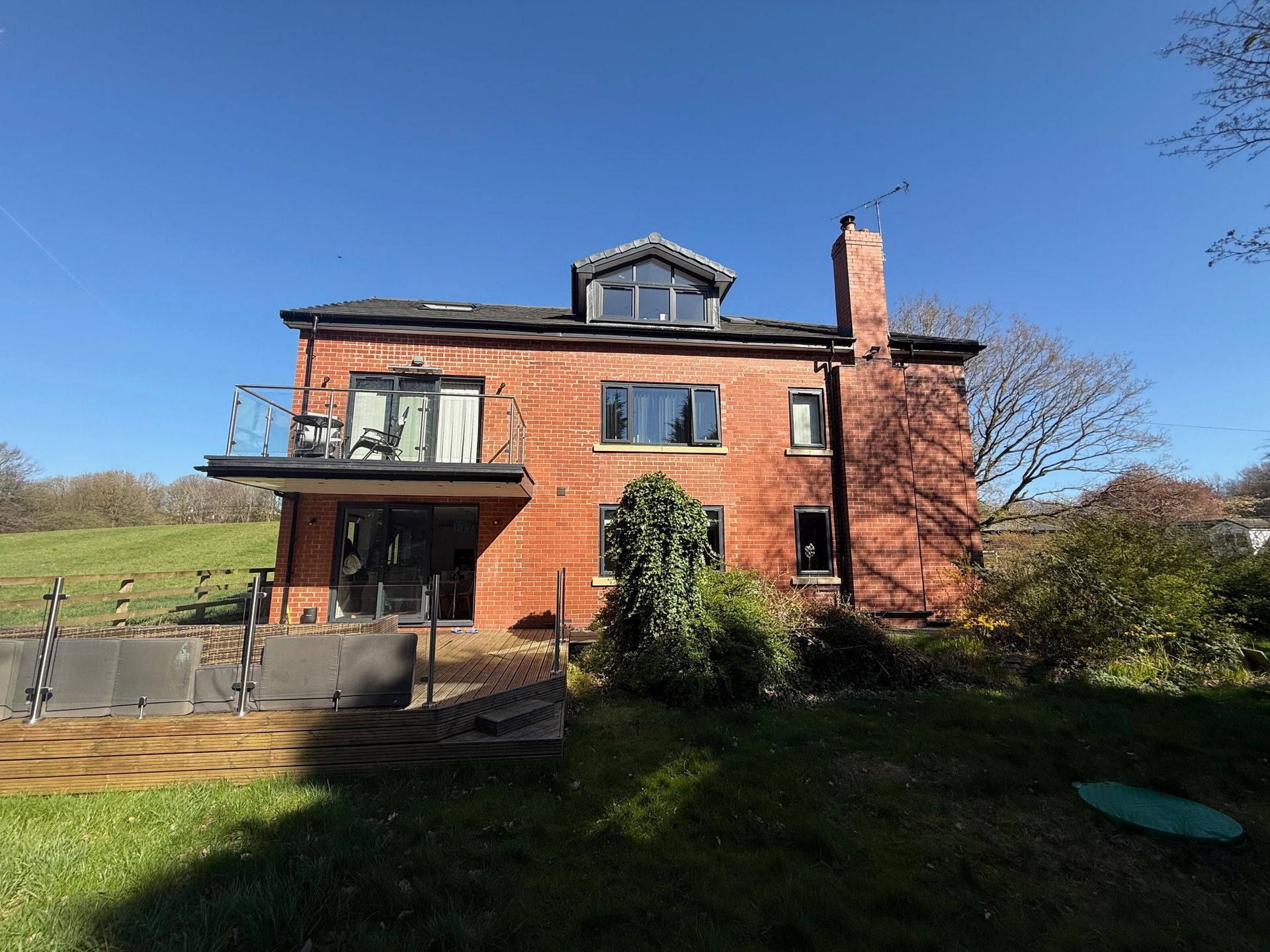 Red brick house with balcony, chimney, and dormer windows, set in a grassy yard under a blue sky.