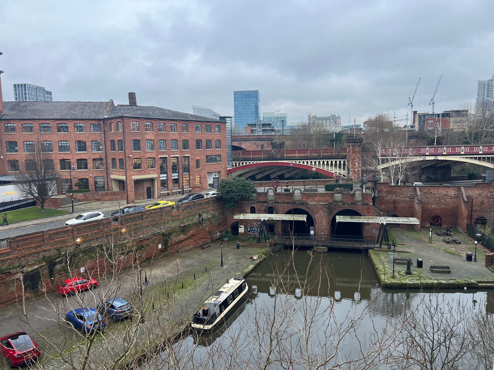 Brick buildings and canal in Manchester, UK, with boats, arches, and a modern skyscraper visible.