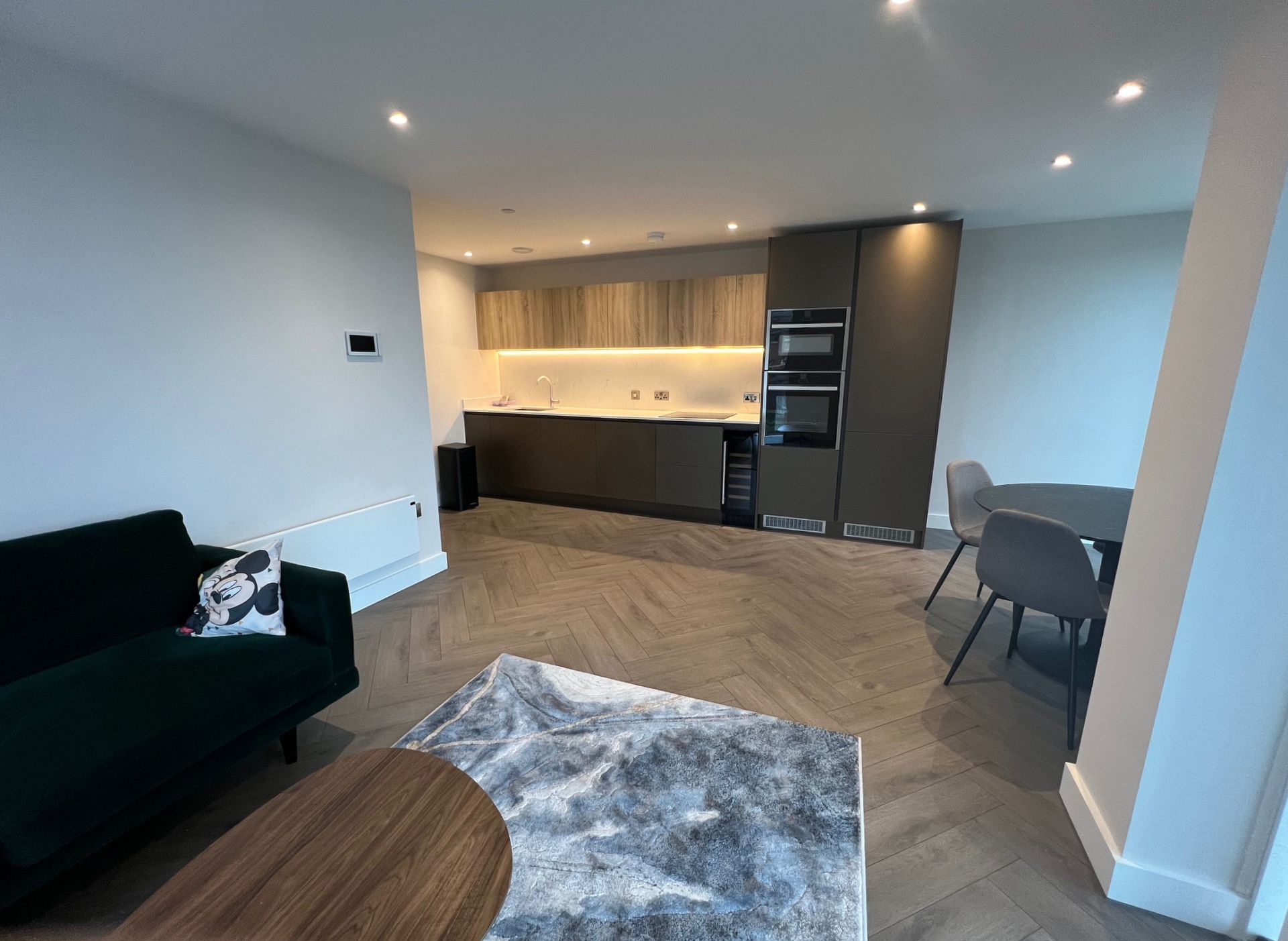 Modern open-plan kitchen and living room with dark green sofa, grey dining table, and light wood cabinets.