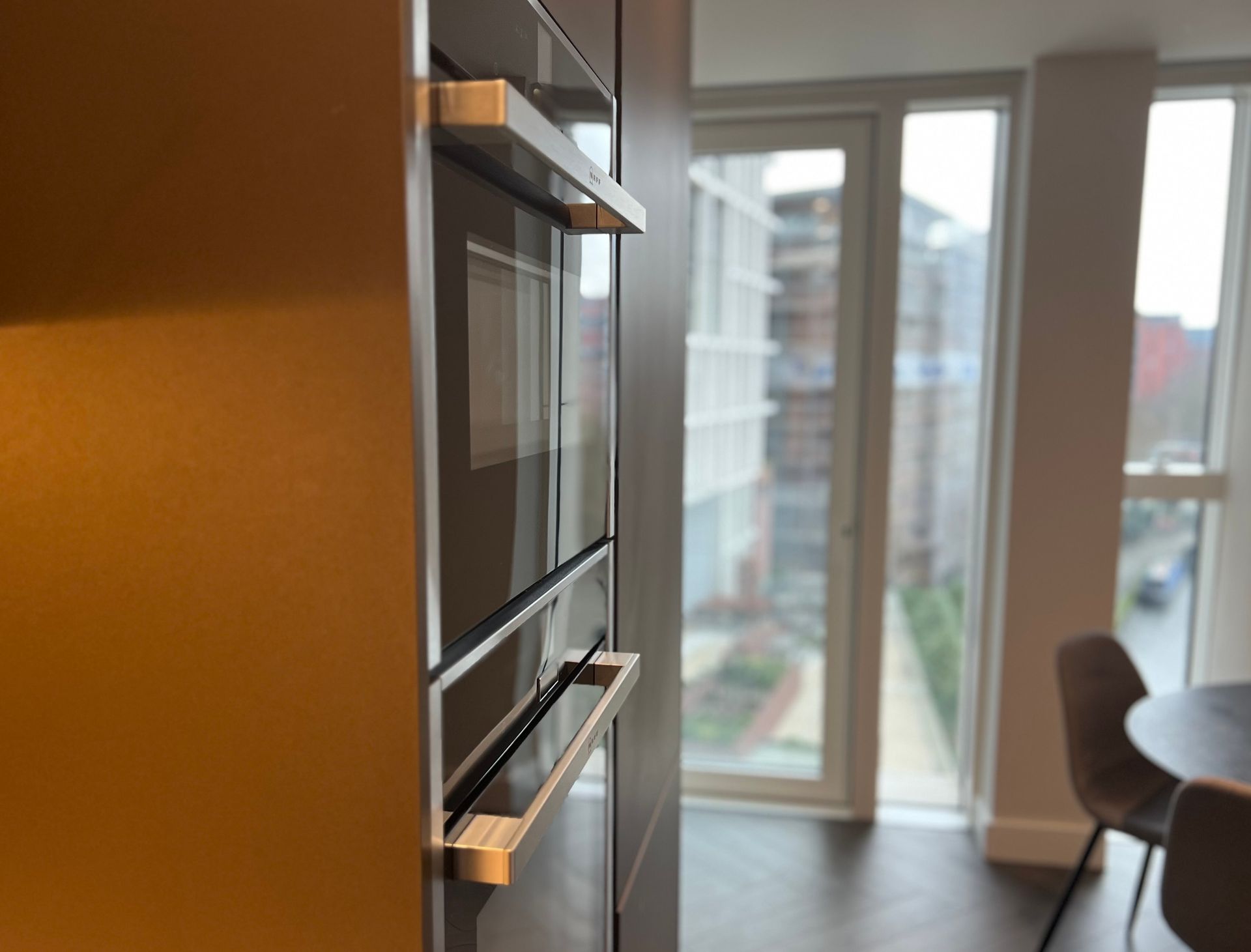 Built-in kitchen appliances with metal handles, next to a window overlooking a cityscape.