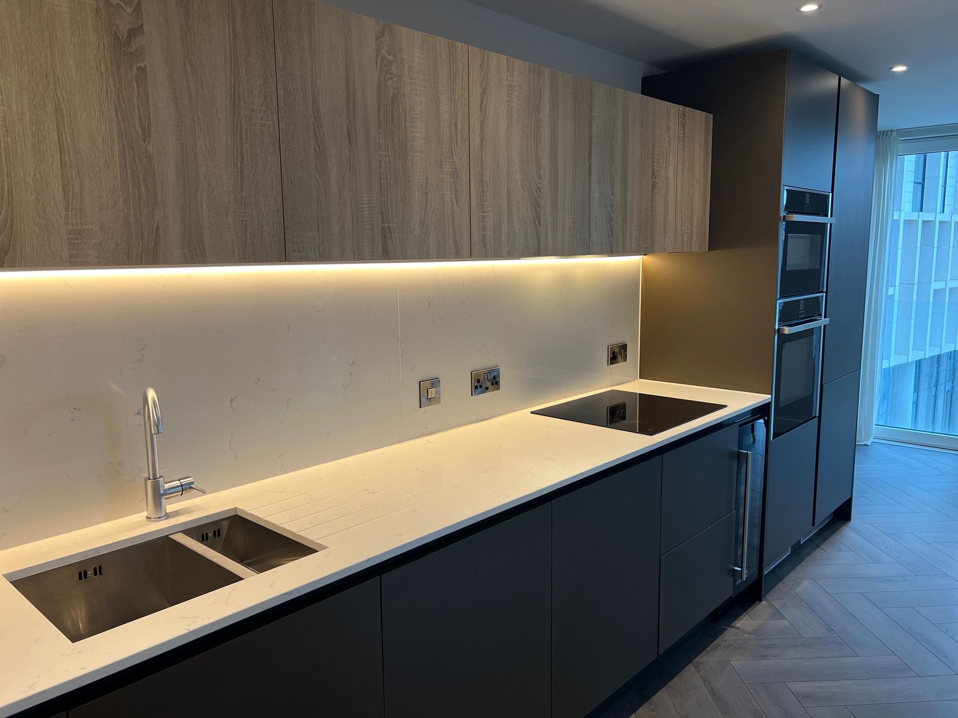 Modern kitchen with dark gray cabinets, light countertops, and wood-toned upper cabinets. Stainless steel sink, cooktop, and built-in appliances.