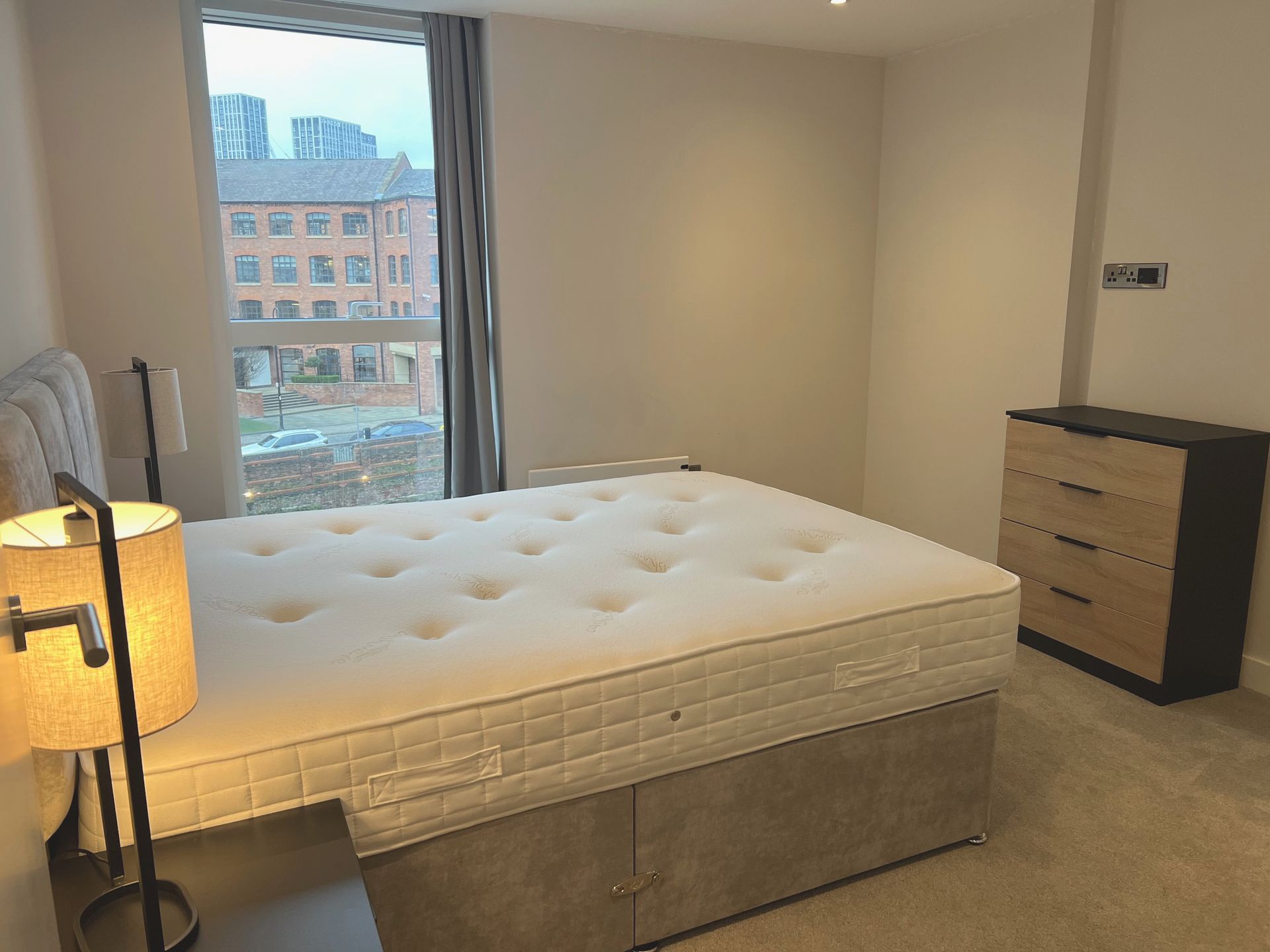 Bedroom with a large bed, nightstand, lamp, and dresser next to a window with a cityscape view.
