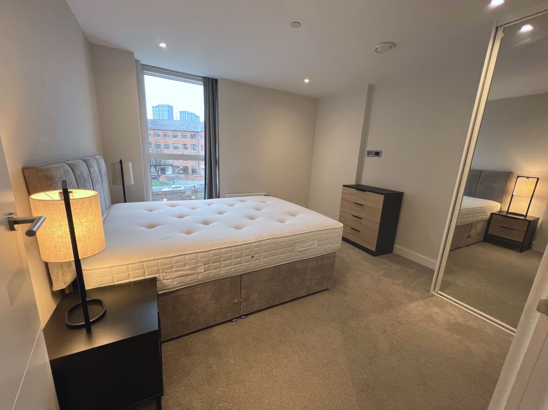 Bedroom with a bed, nightstand, dresser, large mirror, and window with a city view.