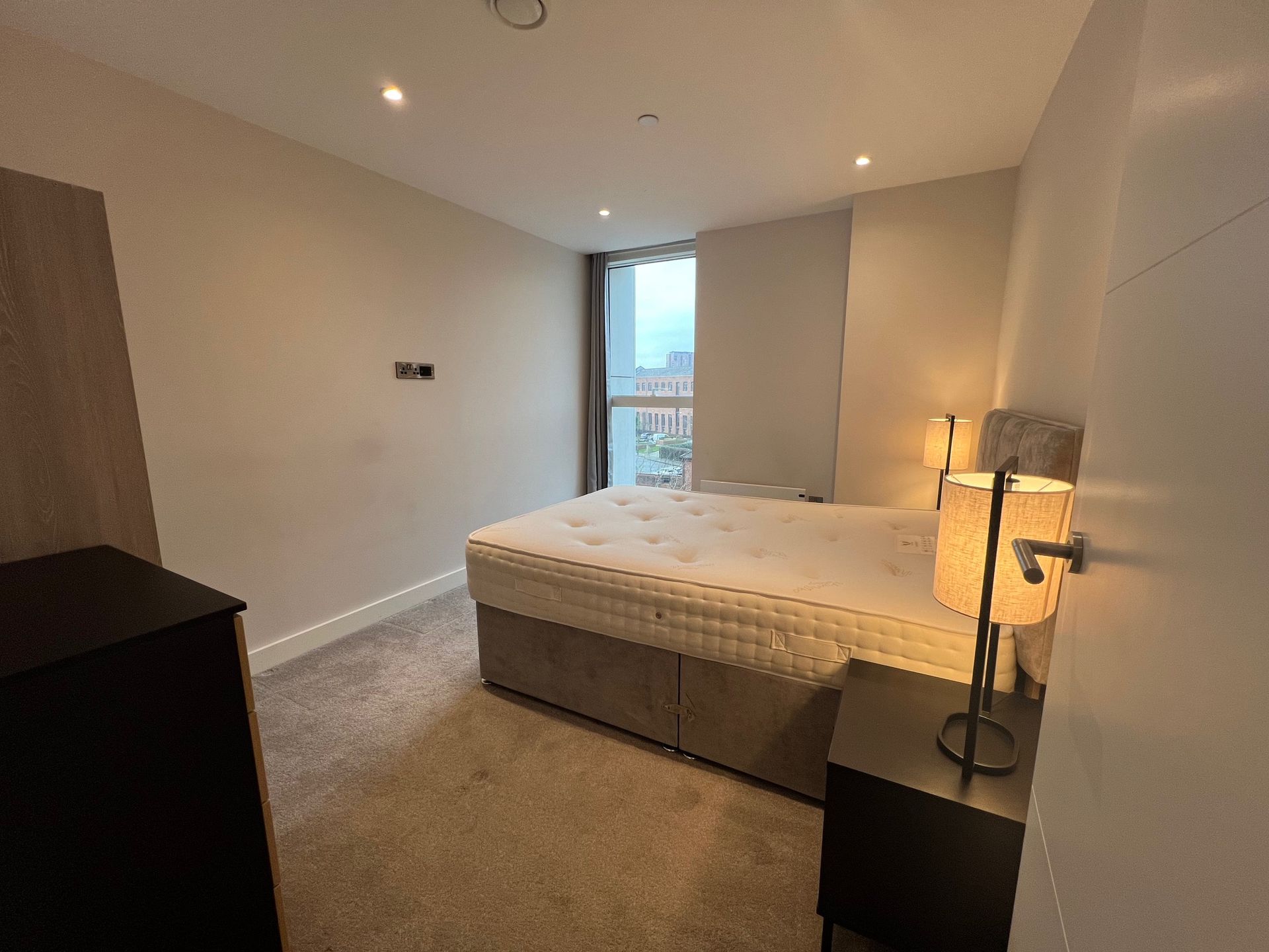 Bedroom with bed, nightstand, lamps, and closet. Window with city view, neutral colors.